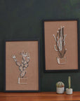 On a white background is a brown piece of wall art with white and black stitching of a cactus in a pot. The art comes in a simple black frame. In this photo, the Prickly Pear wall art is staged next to another piece of wall art. 