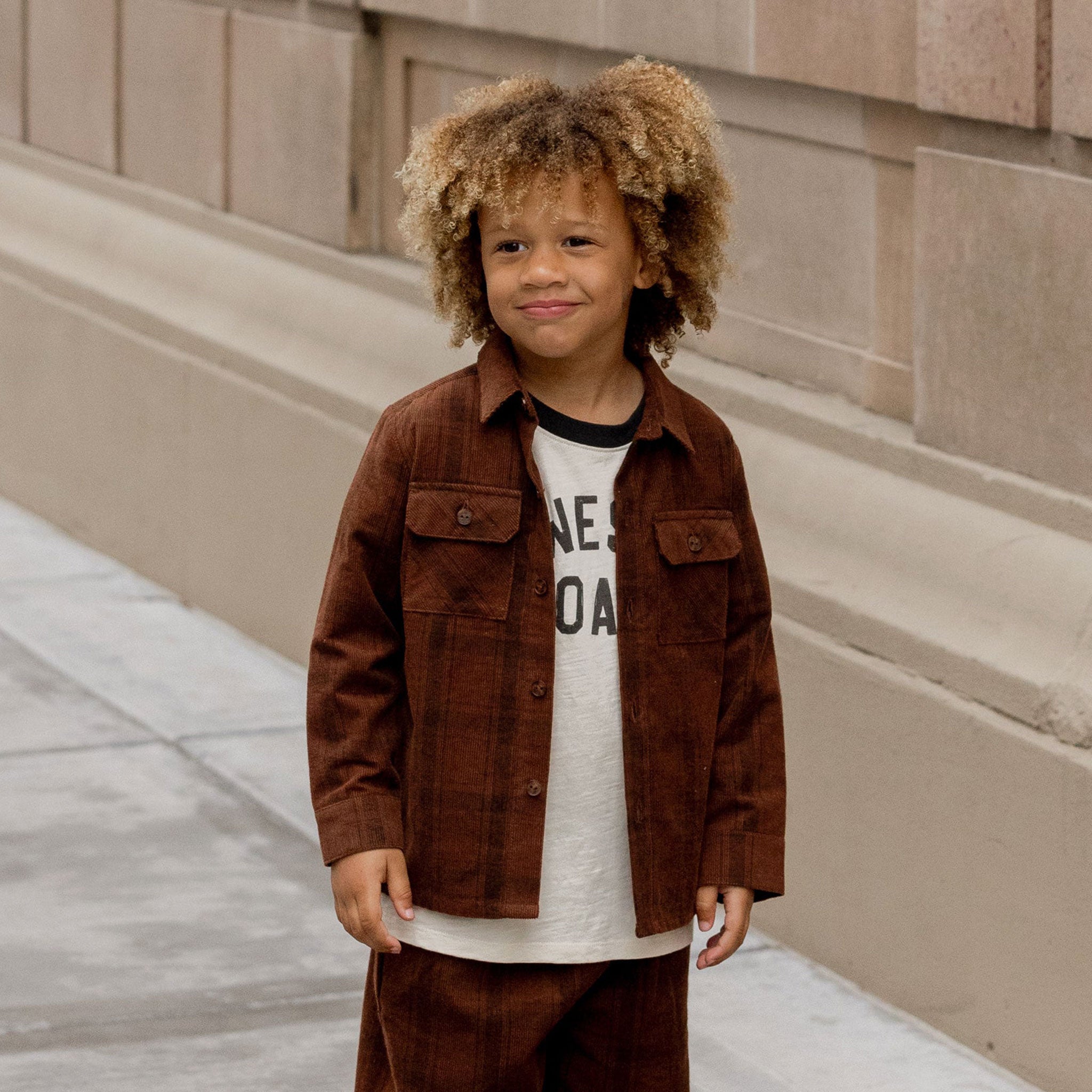 Little boy wearing a brown plaid long sleeve shirt. The shirt has two pockets and buttons down the front
