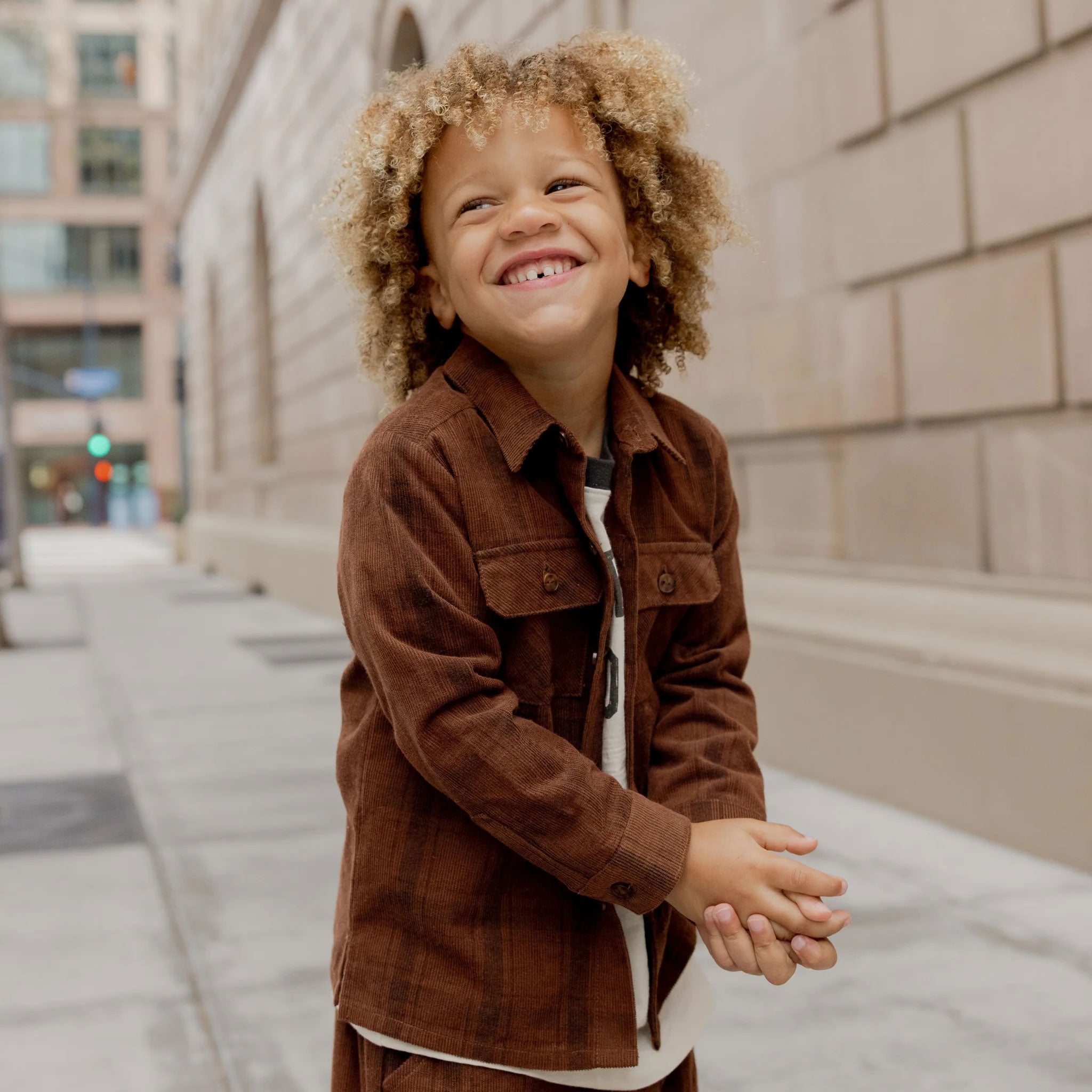 Little boy wearing a brown plaid long sleeve shirt. The shirt has two pockets. 