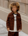 Little boy wearing a brown plaid long sleeve shirt. The shirt has two pockets and buttons down the front
