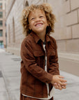 Little boy wearing a brown plaid long sleeve shirt. The shirt has two pockets. 