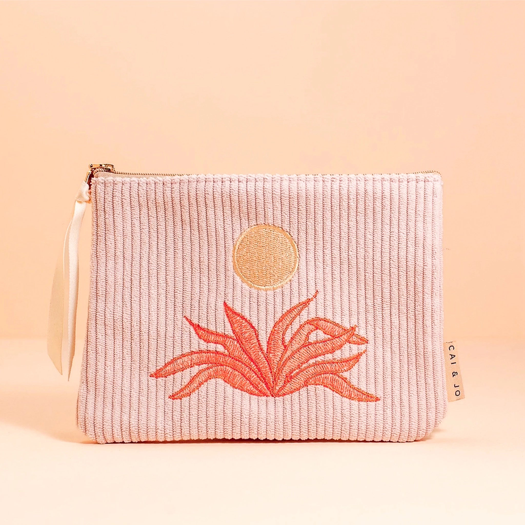 A light pink corduroy pouch with a sun and an orange aloe plant graphic in the center. The pouch has a single zipper going across the top.
