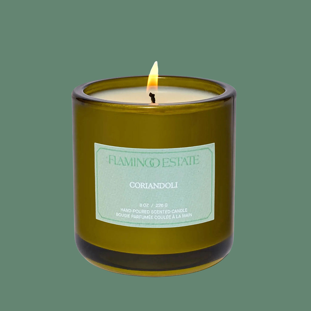 A green glass candle with a white label that reads, 'Flamingo Estate Coriandoli'. 
