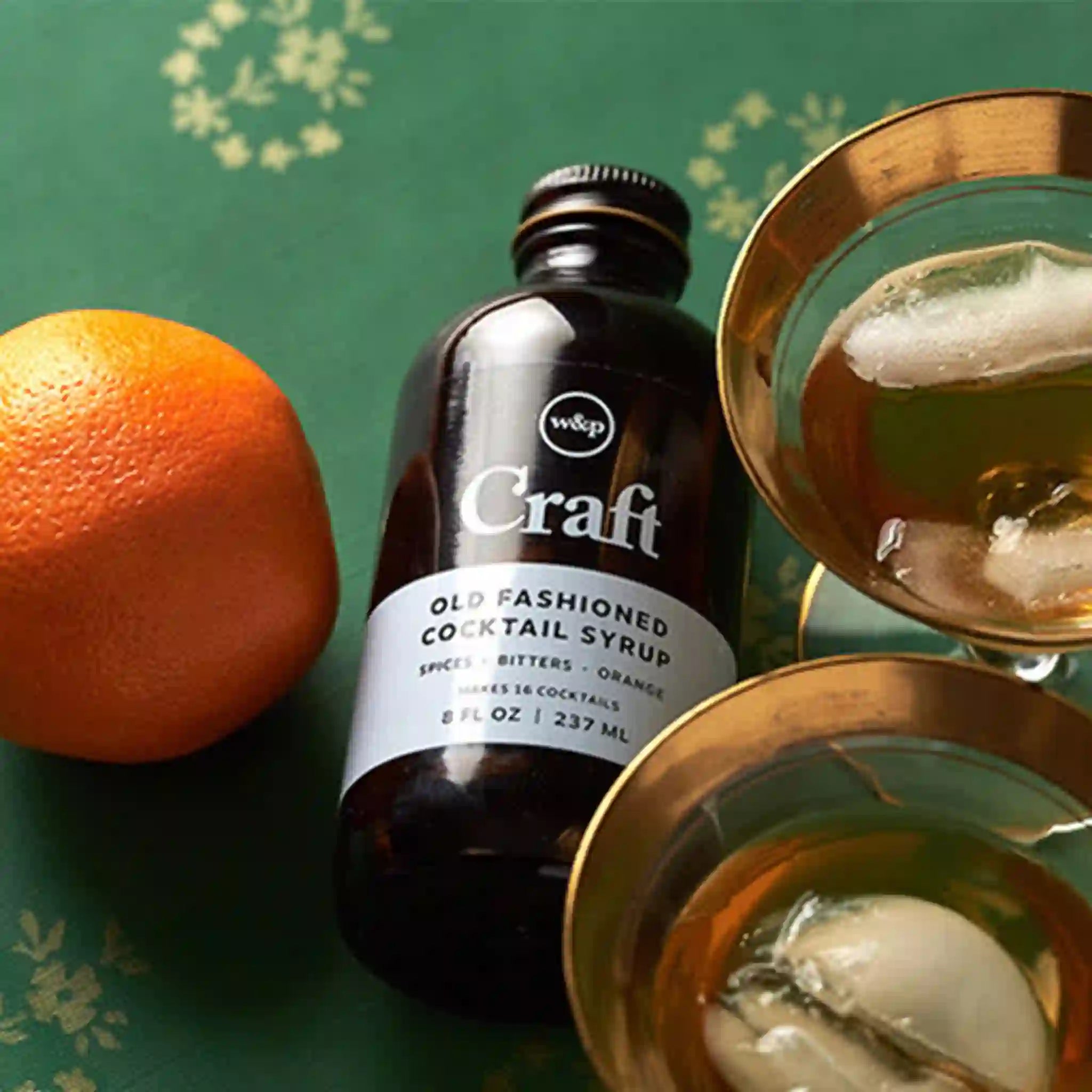 On a green background is a glass bottle of cocktail syrup. 