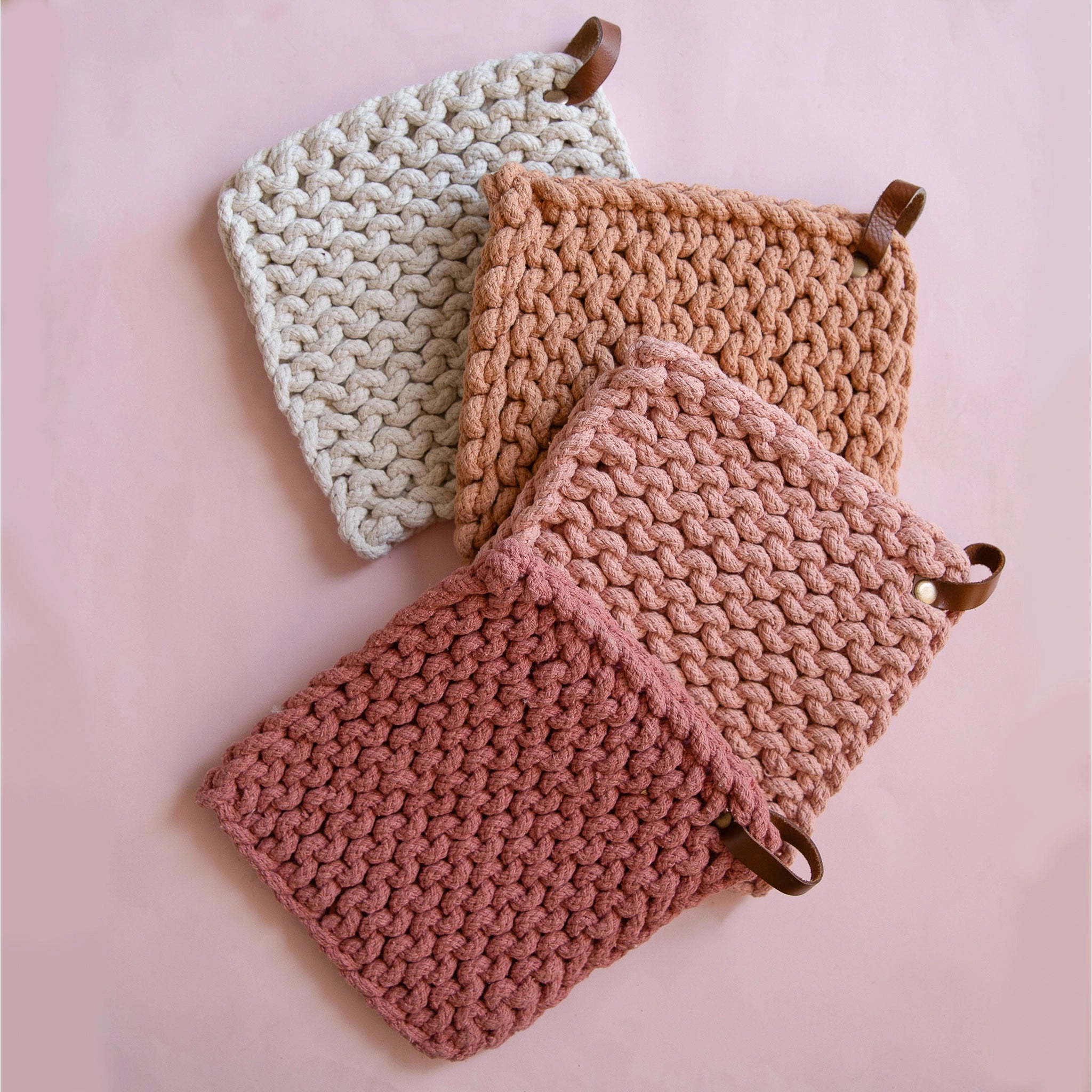 On a pink background is four crocheted pot holders in four different shades. There is an ivory, two pink shades, and a terracotta shade.