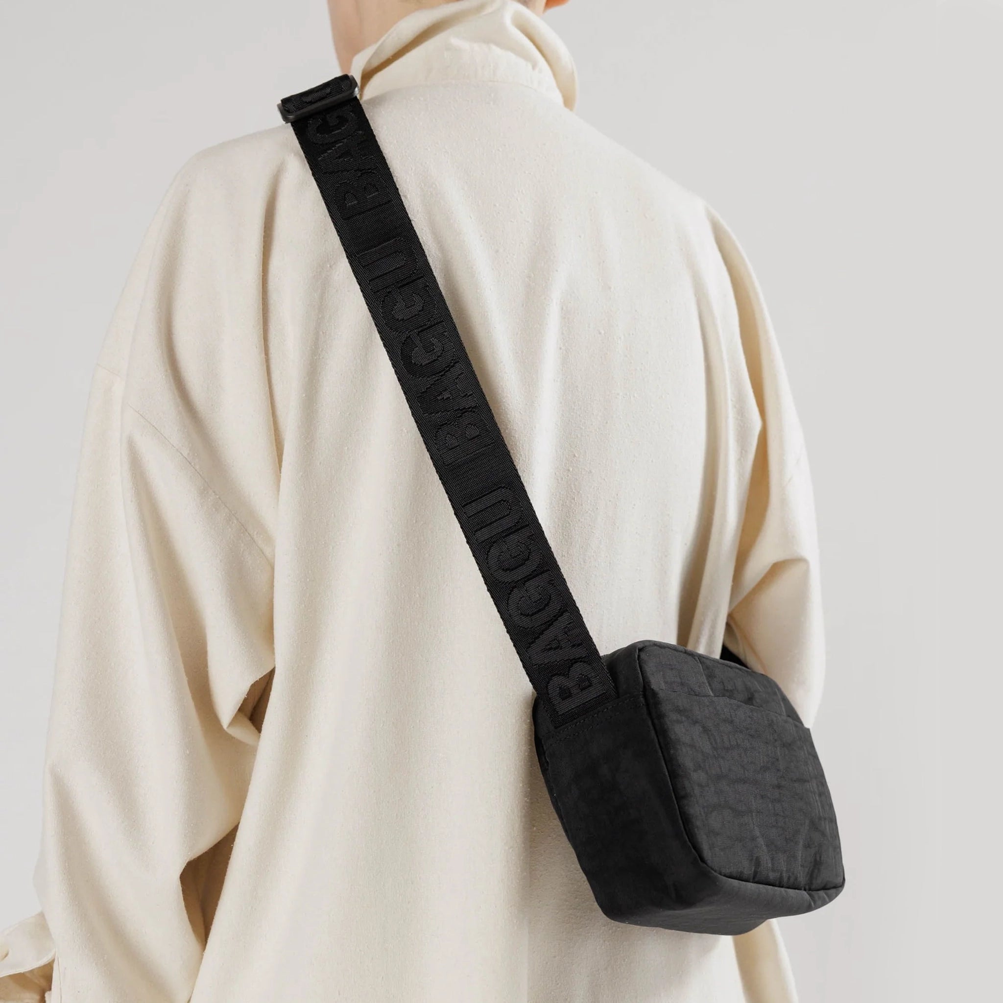 On a light grey background is a model wearing a black crossbody made of sturdy nylon material with a black adjustable strap that has "BAGGU" stitched into it in a shinier black material. 