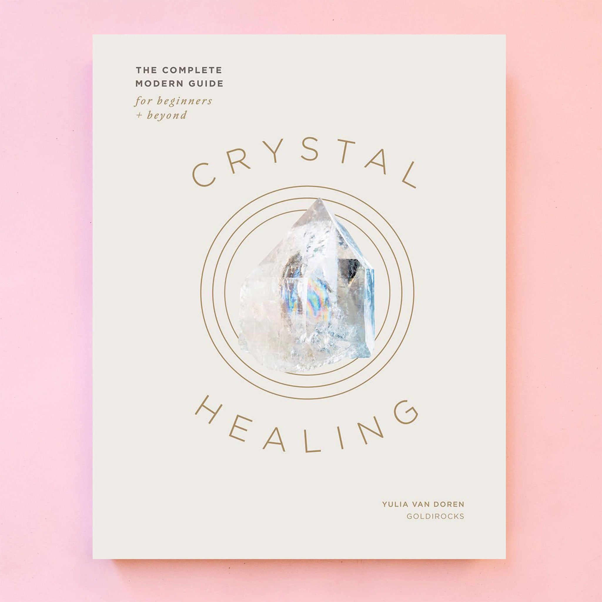 On a pink background is a white book with a crystal in the center and the title around it reading, &quot;Crystal Healing&quot;. 