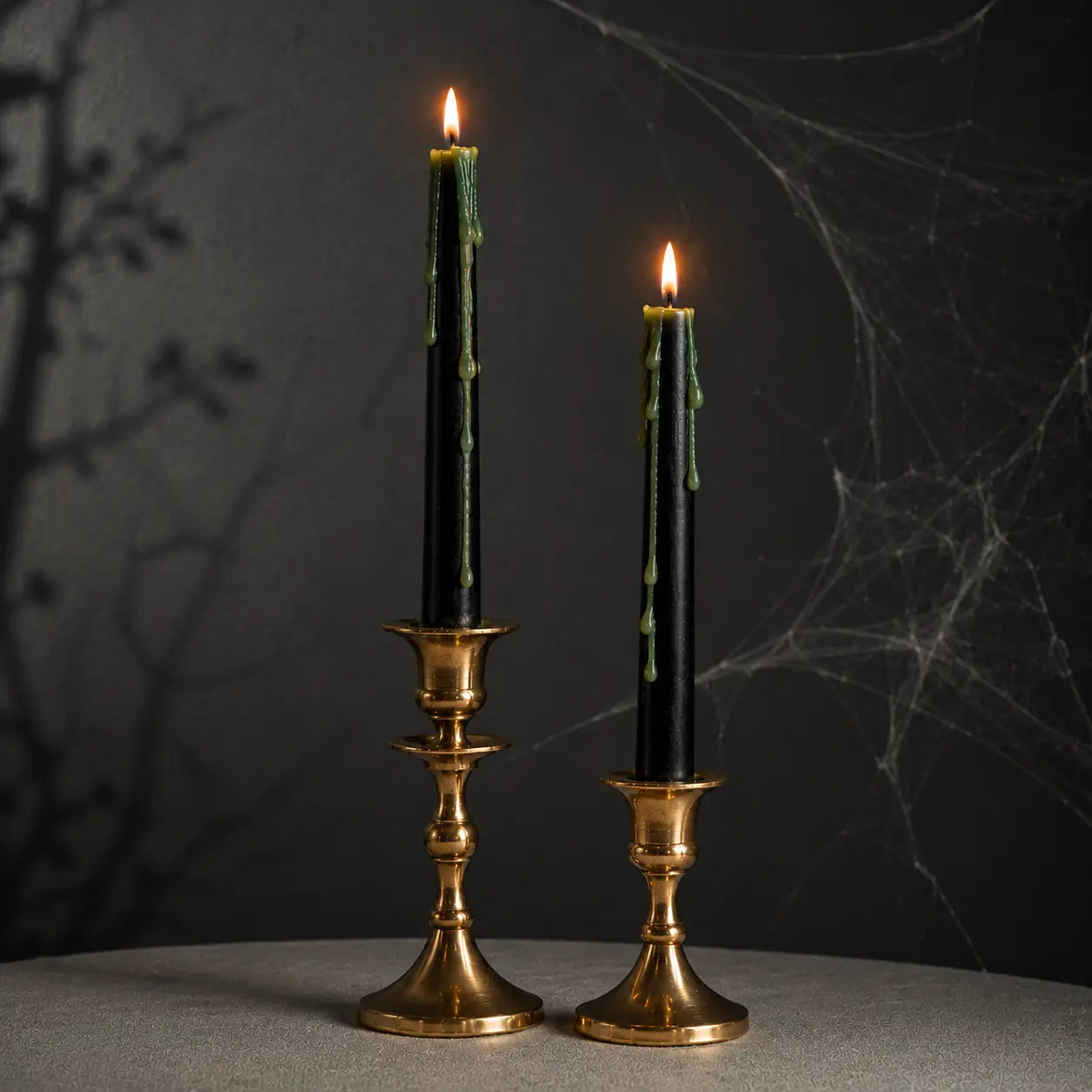 Two black taper candles with green wax drips in ornate gold candlesticks, lit and set against a dark gothic background with spiderwebs and tree shadows.