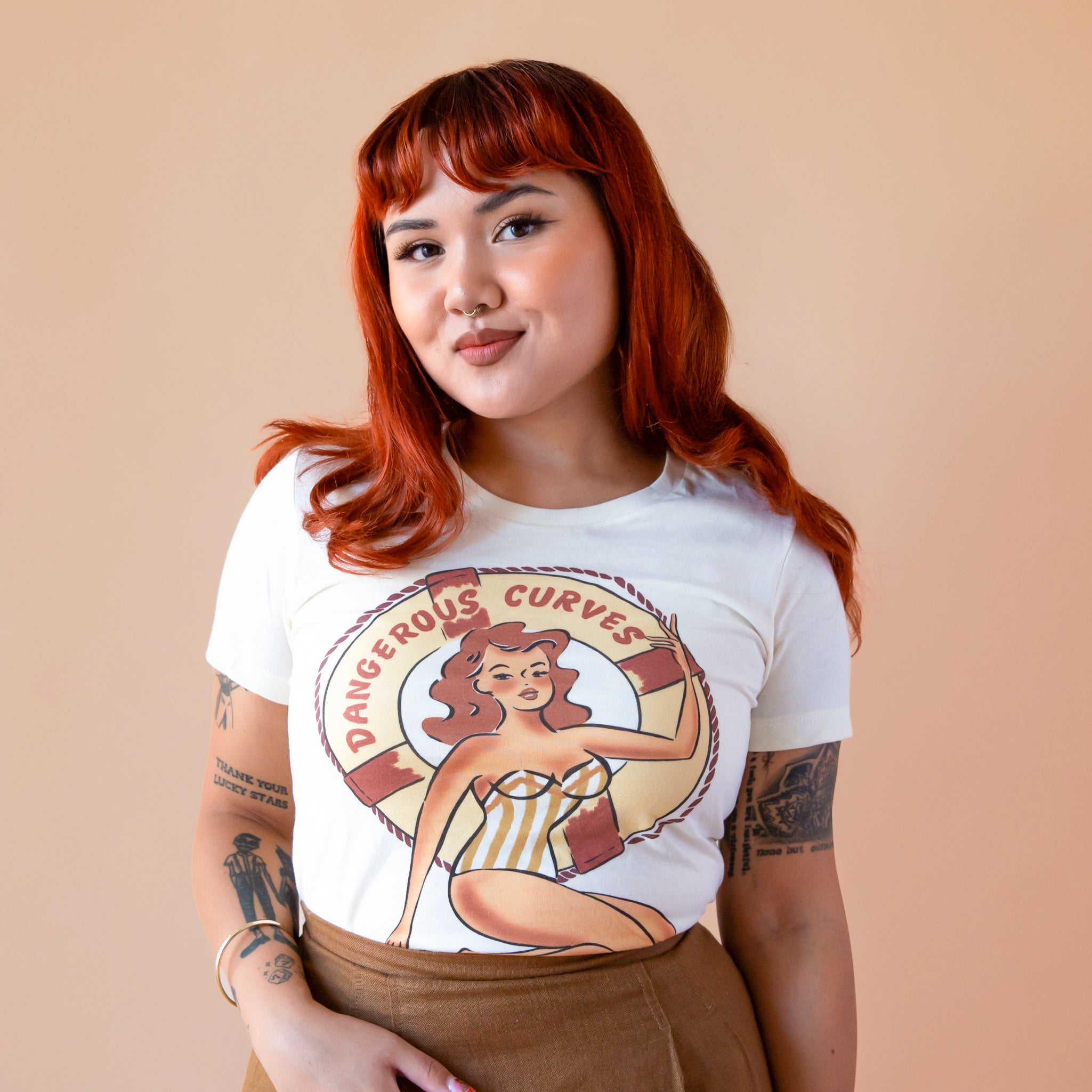 A model with red hair wearing an ivory tshirt with a design of a graphic of a woman with a swimsuit in front of a life raft that reads, 'Dangerous Curves'. 