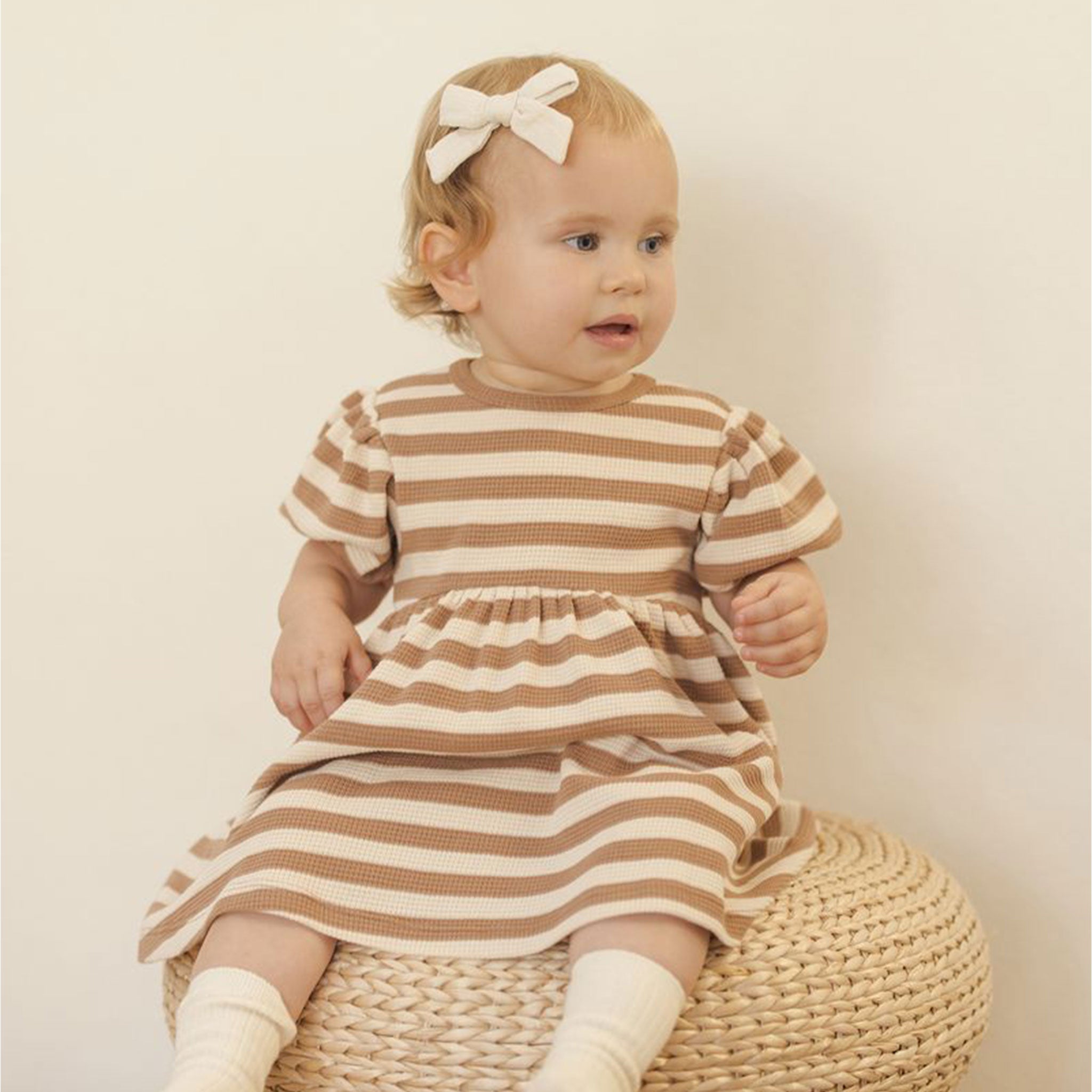 Baby wearing a waffle knit kids dress. The dress has bubble sleeves and a brown/off white stripe pattern. She has a bow in her hair and socks on. 