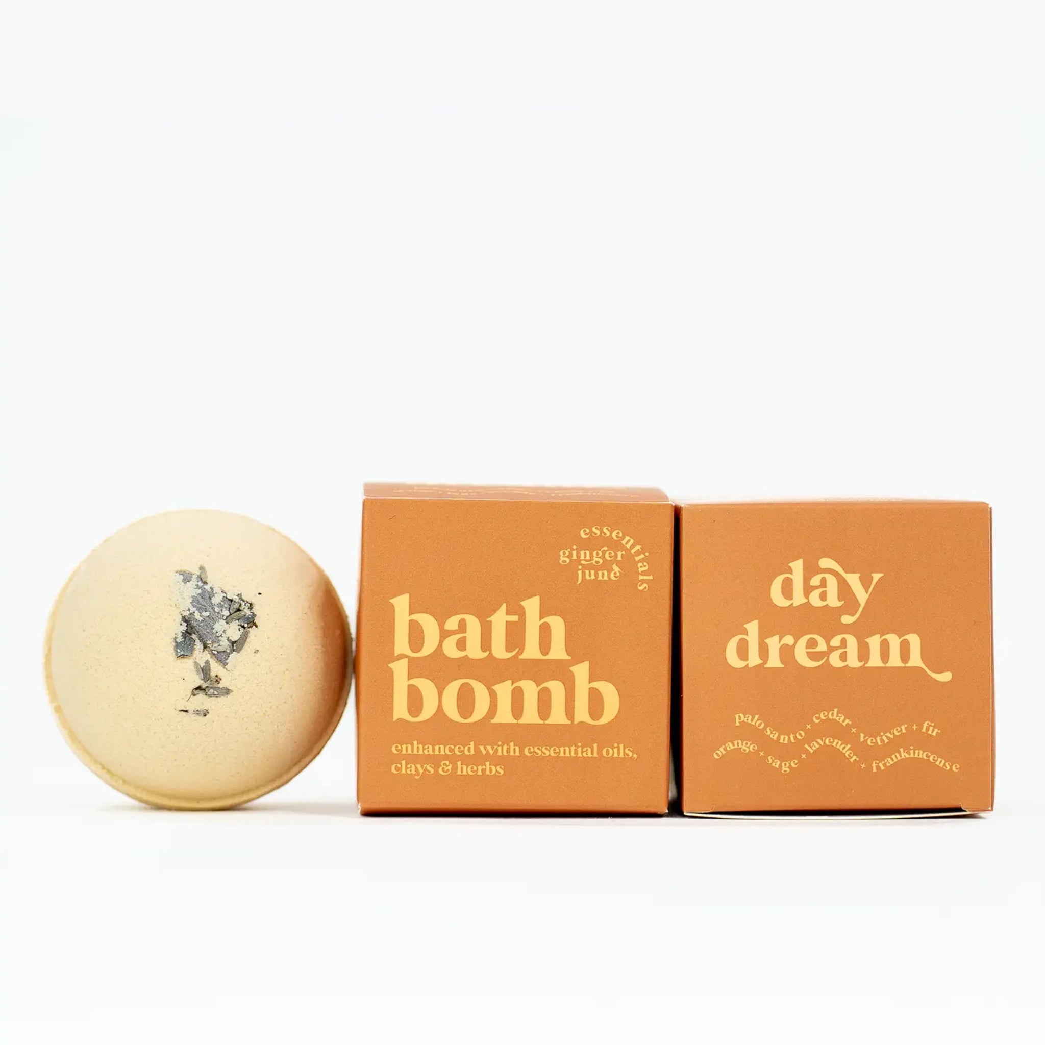 On a neutral background is a round bath bomb with an orange hue and a orange box with yellow text that reads, &quot;bath bomb enhanced with essential oils, clays &amp; herbs&quot;. 