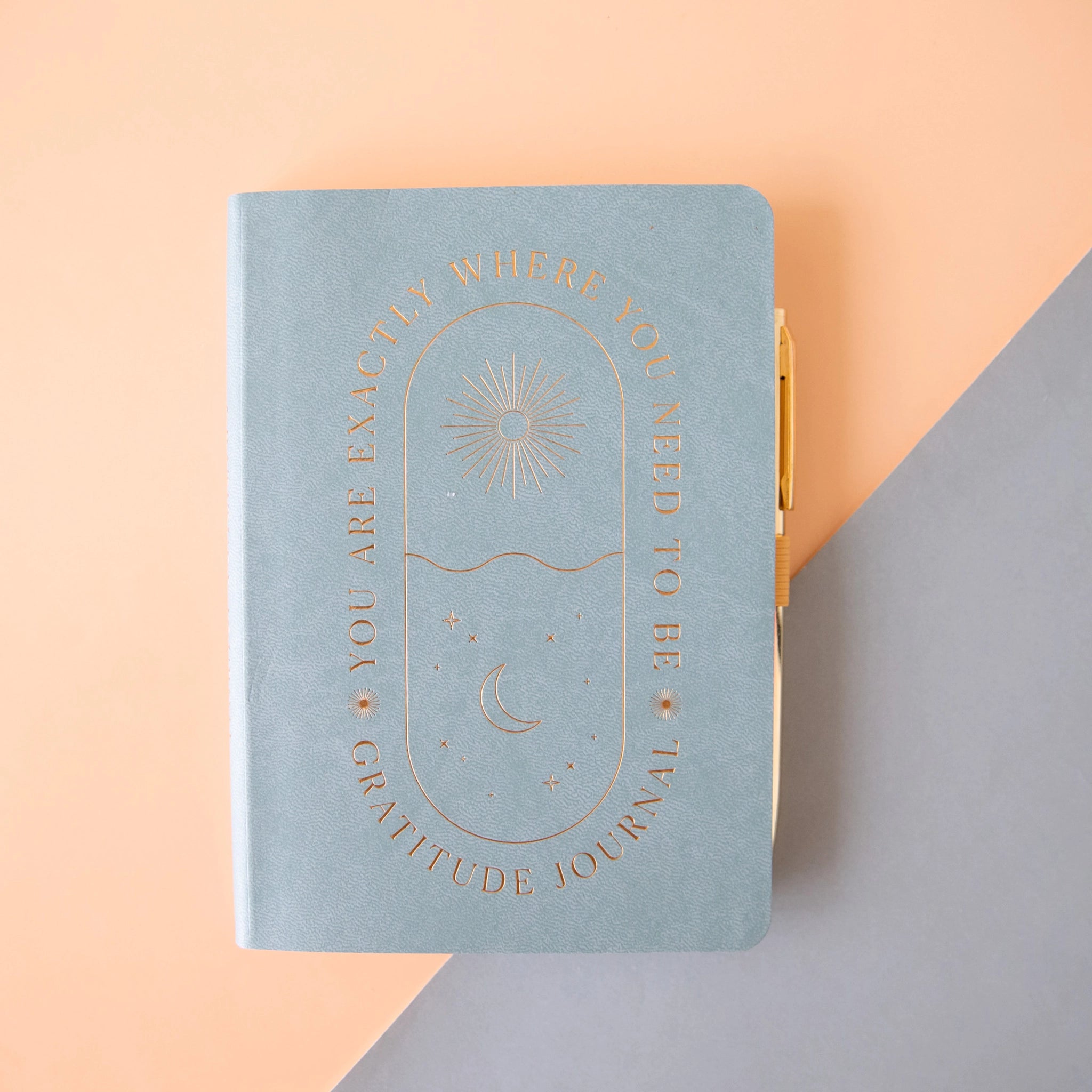 On a peach and light blue background is a light blue journal with gold text that reads, "You Are Exactly Where You Need To Be" and "Gratitude Journal" around an oval shape that has a sun and a moon inside. It also includes a gold pen. 