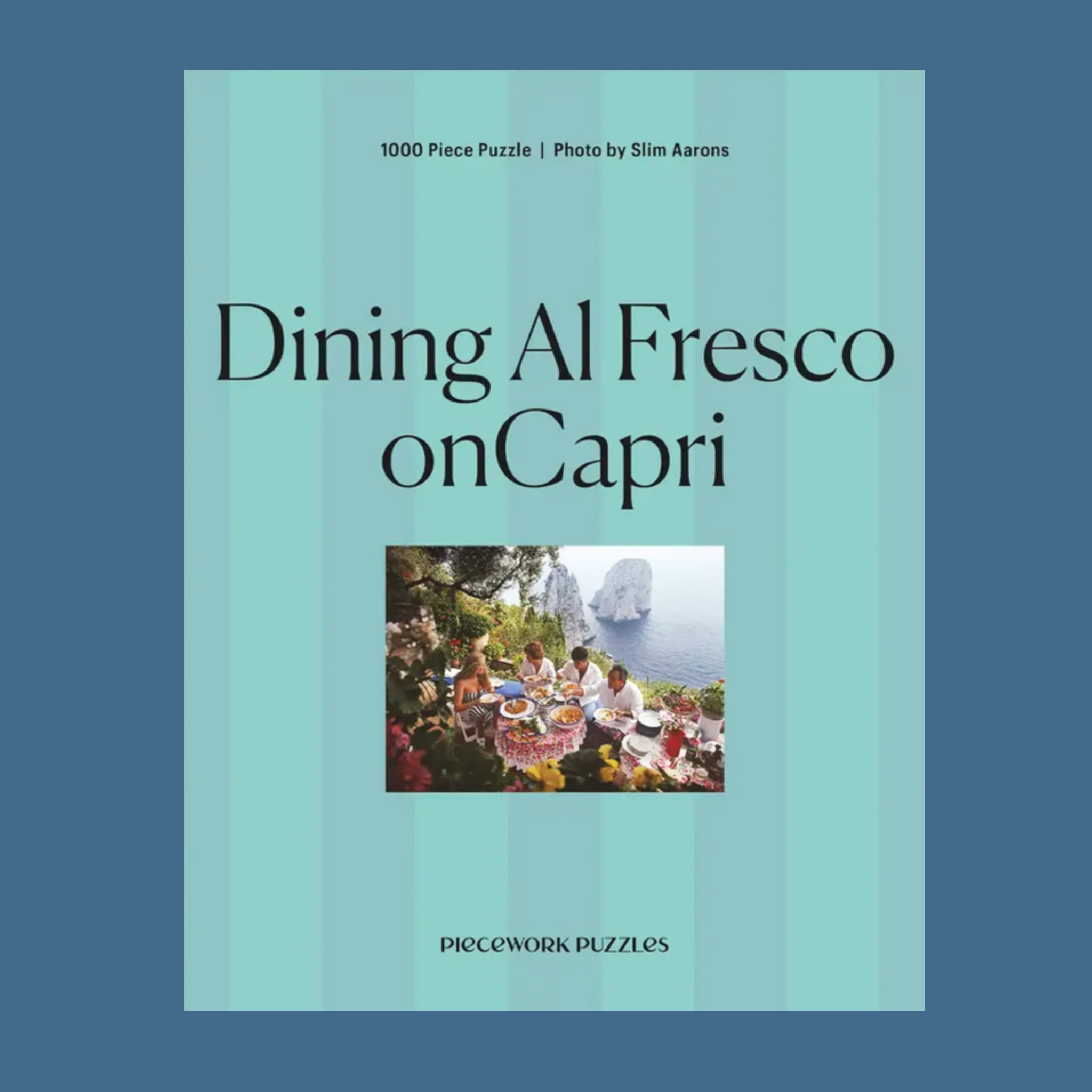 A blue box of a puzzle with a photo of people gathered at a family style table eating a spread of Italian food overlooking the coastline. 