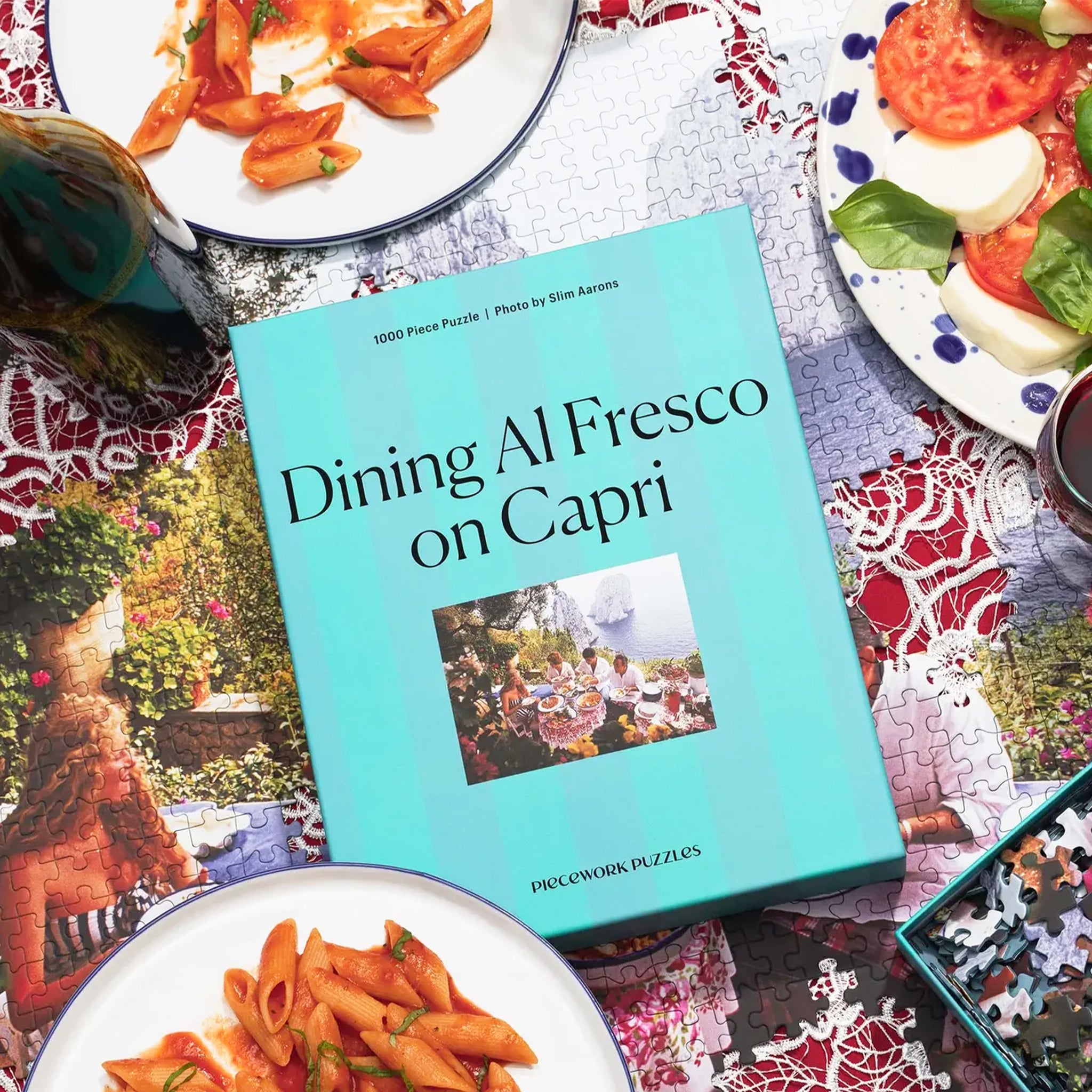 A blue box of a puzzle with a photo of people gathered at a family style table eating a spread of Italian food overlooking the coastline. 