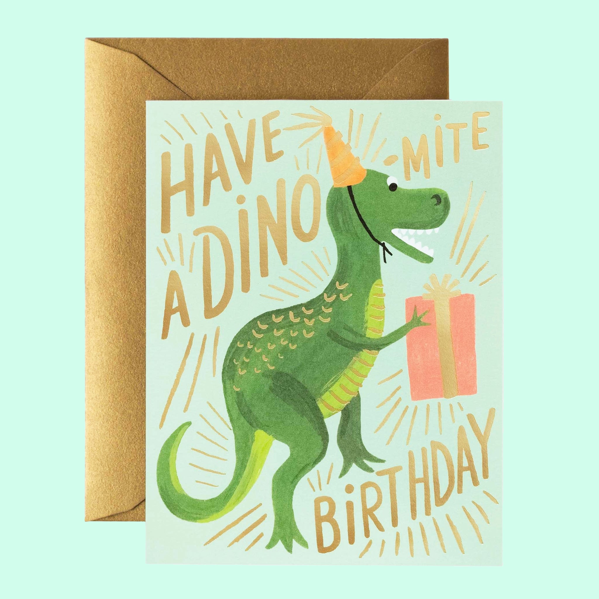 A mint card with gold letters that read, "Have A Dino-Mite Birthday" with a illustration of a green t-rex dinosaur wearing a yellow striped birthday hat and holding a pink birthday gift as well as a coordinating envelope.