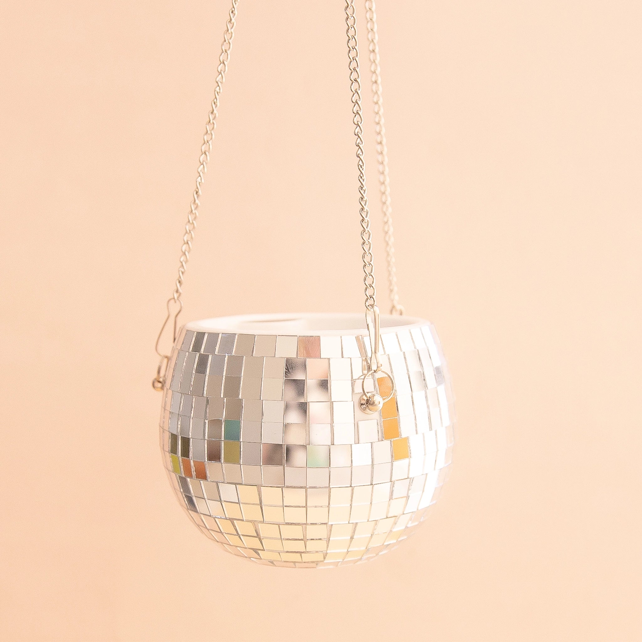 On a peachy background is a silver mirrored glass disco ball shaped planter with a silver chain hanger. 