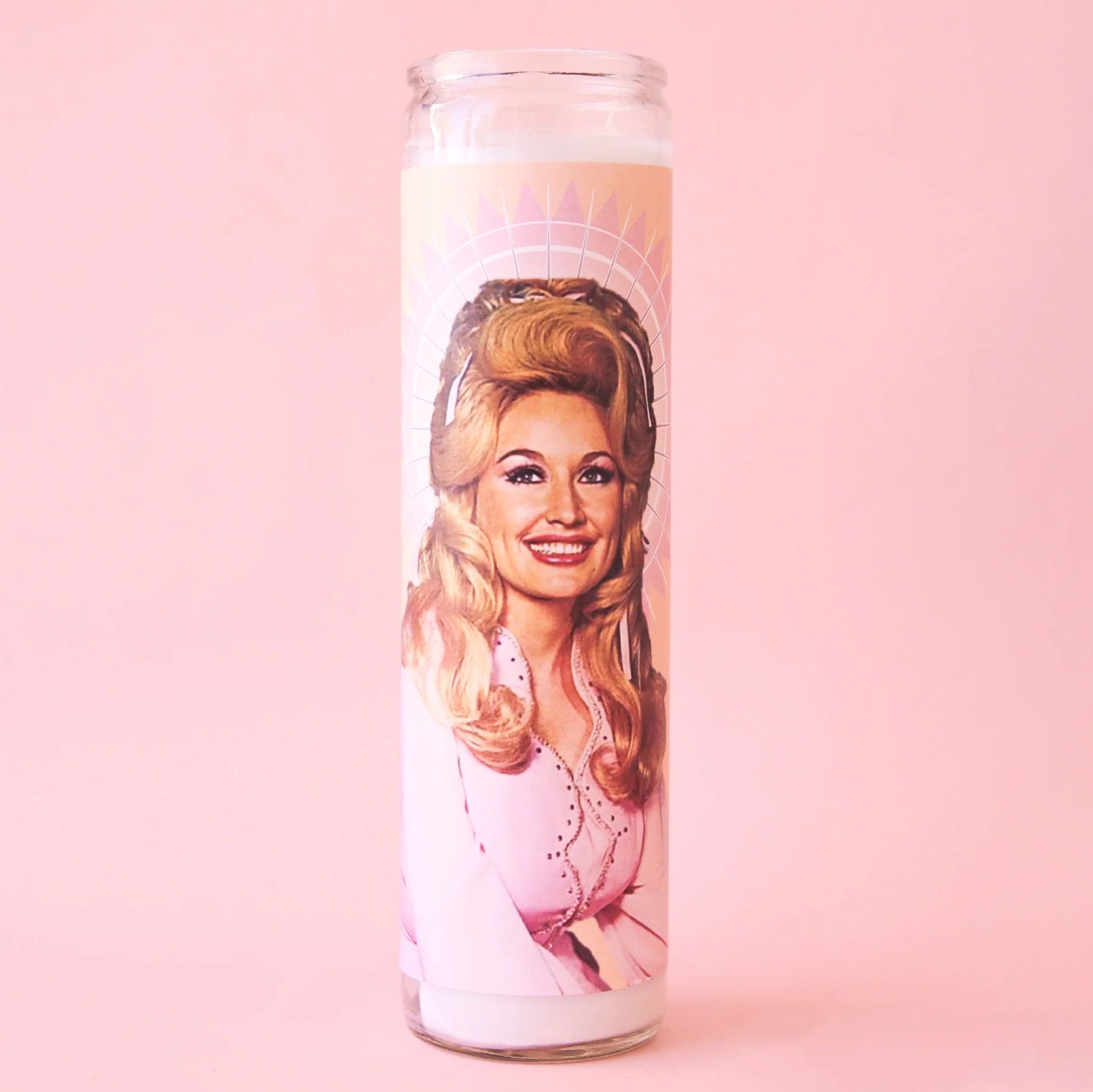A tall glass prayer candle. On the front is a picture of Dolly Parton with long blonde hair and a pink jacket. 