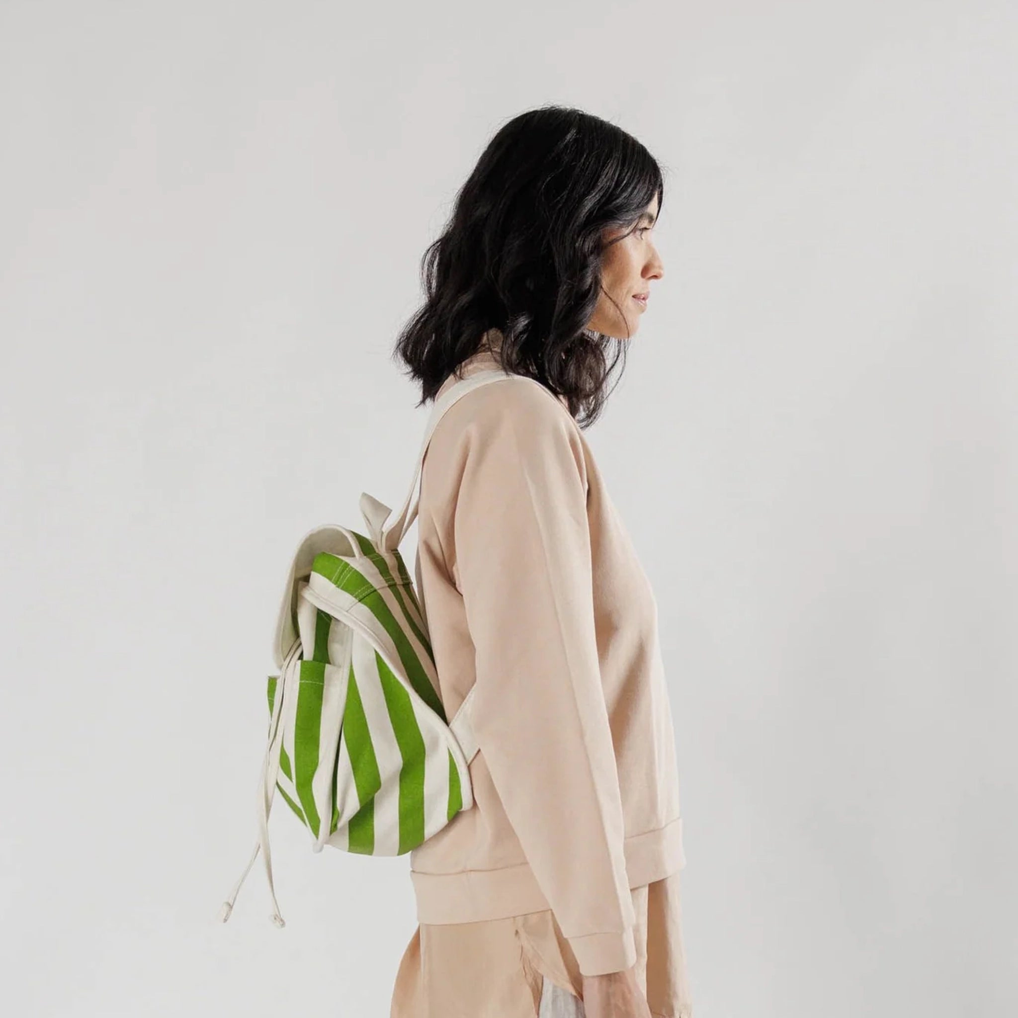 A model wearing a green and white striped backpack with a folded flap and a drawstring opening along with two exterior pockets on the outside.