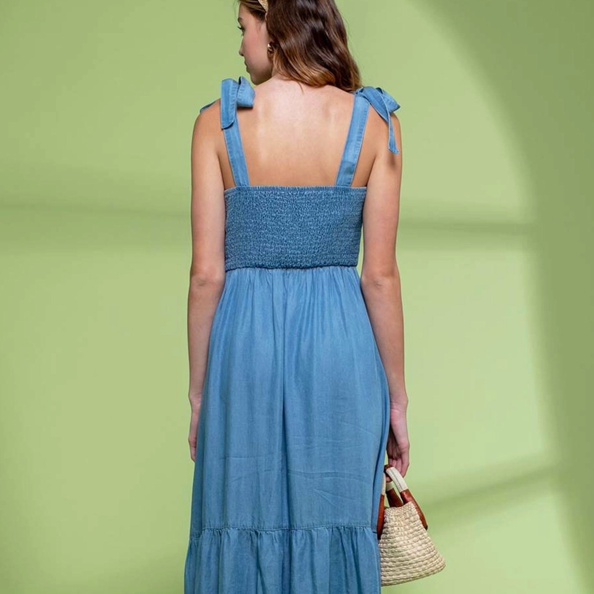 On a green shadow background is a model wearing a blue flowy midi dress with tie shoulder straps and a square neckline.