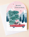 On a neutral background is a pink arched card with a snowy mountain background with a red convertible stacked with Christmas trees. 