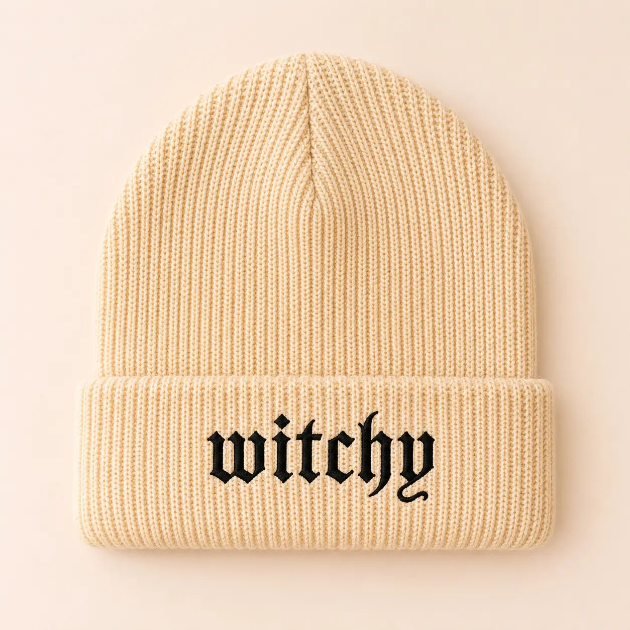 A cream-colored ribbed knit beanie hat with the word 'witchy' embroidered in a black gothic font on the front cuff, set against a solid pale peach background.