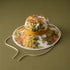 A wide-brim sun hat with a white base and a dense yellow, orange, and green floral print, featuring a mustard yellow band and white chin straps, set against a solid olive green background.
