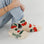 On a white background is a model wearing cream colored socks with a bright red cherries design on it along with "Baggu" on the bottom of the foot. 