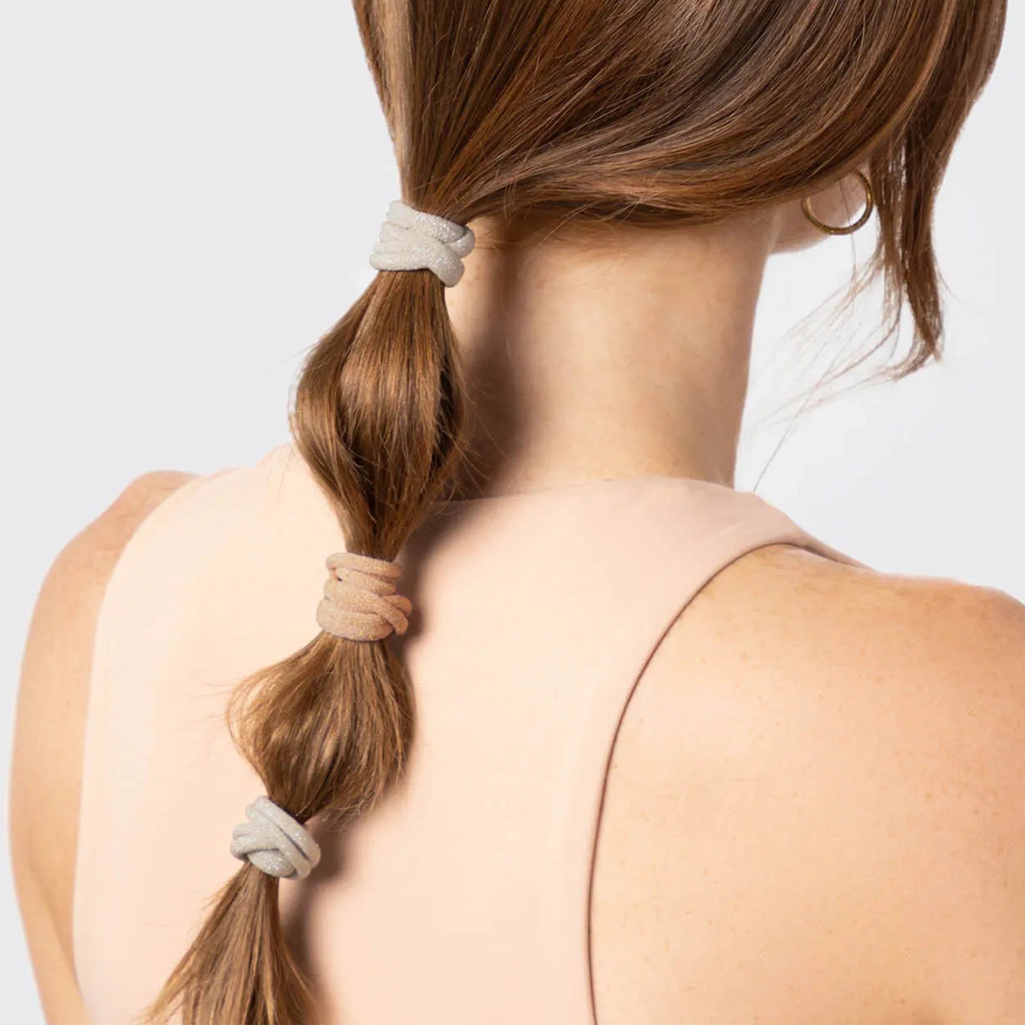 A model wearing neutral elastic hair ties with a glitter finish. The colors range from a neutral tans, brown, and grey.