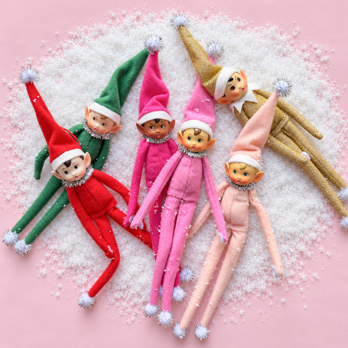 elf ornaments in a dogpile on fake snow and a pink background.