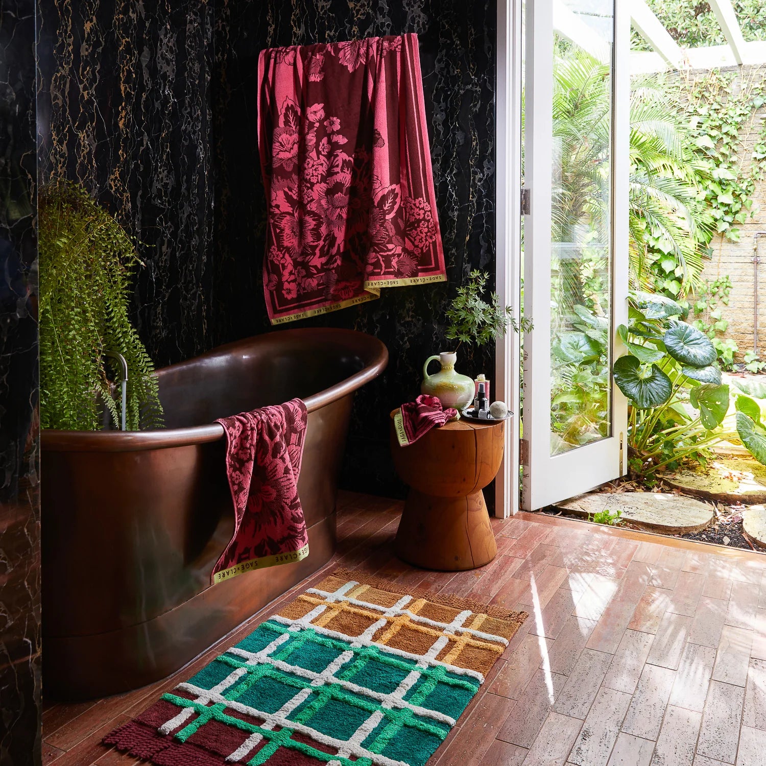 Bathroom with a copper soaking tub and dark marble wall, featuring burgundy floral towels draped over the tub and hanging above, with greenery and an open door to a garden.