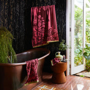 Bathroom with a copper soaking tub and dark marble wall, featuring burgundy floral towels draped over the tub and hanging above, with greenery and an open door to a garden.