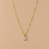 A delicate gold chain necklace with a small teardrop-shaped pendant set with a clear marquise-cut gemstone, displayed against a solid beige background.