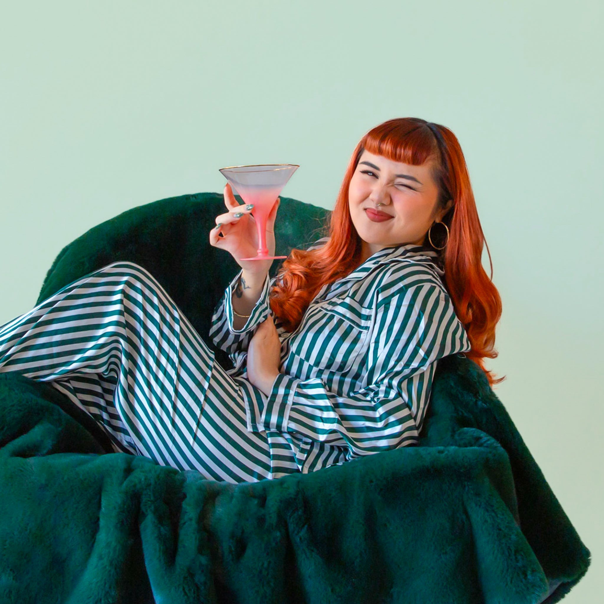 A person with long red hair lounges in a plush dark-green chair, wearing green-and-white striped pajamas and holding a pink cocktail glass while giving a playful wink.