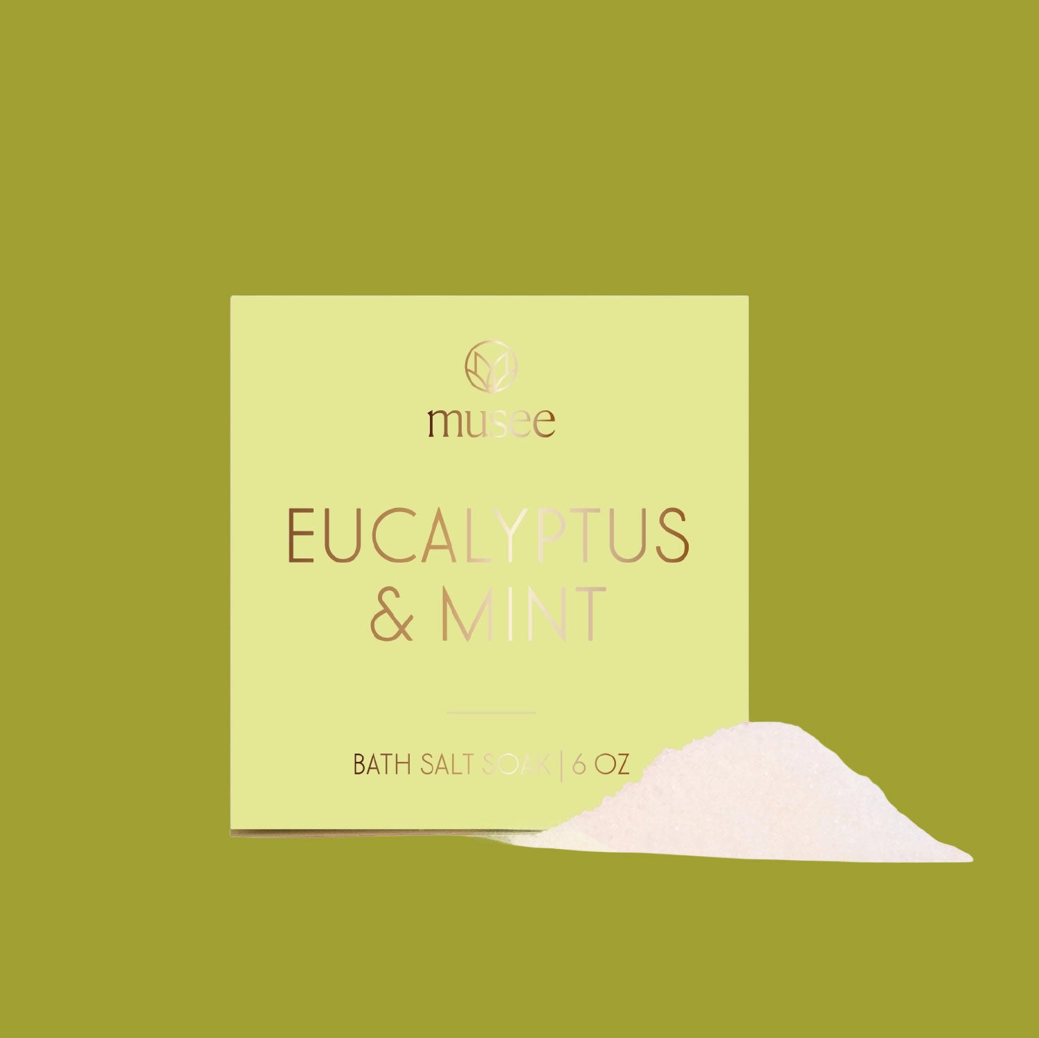 A pale green box labeled “Musee Eucalyptus & Mint Bath Salt Soak” sits against an olive-green background. A small mound of white bath salts is poured out in front of the package.