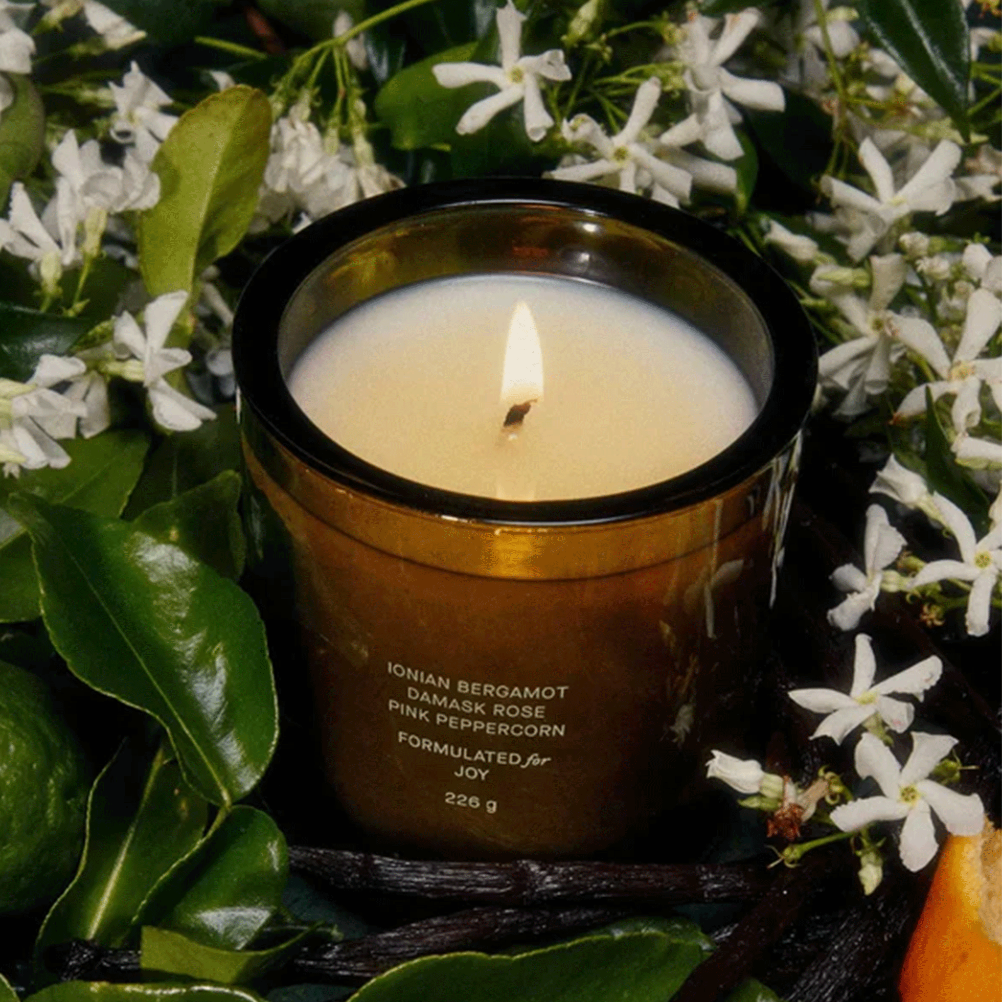 A glass candle with white wax and small text that reads, &quot;Ionian Bergamot Damask Rose Pink Peppercorn Formulated for Joy&quot;.
