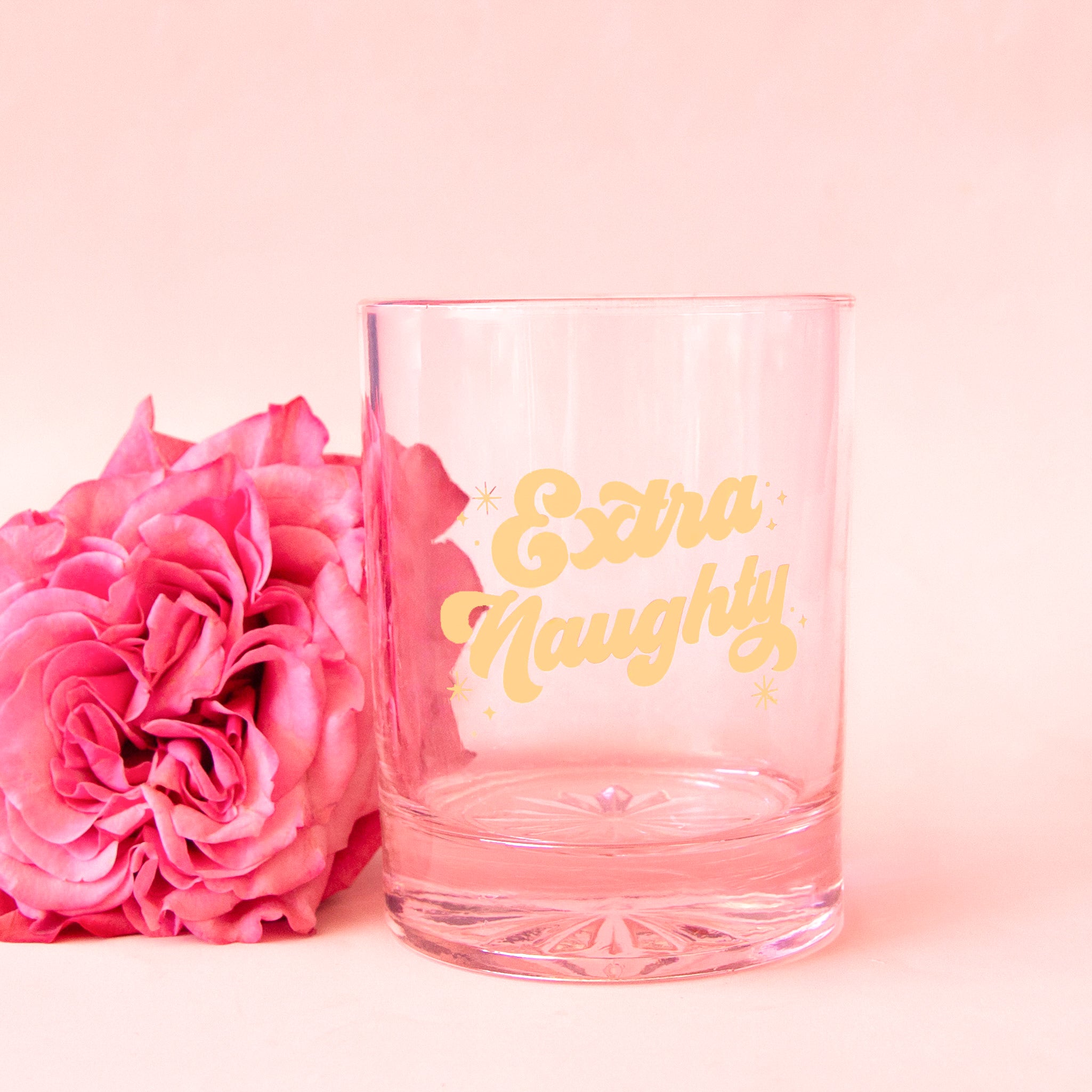 A light pink, clear tumbler glass with gold text that reads, "Extra Naughty".
