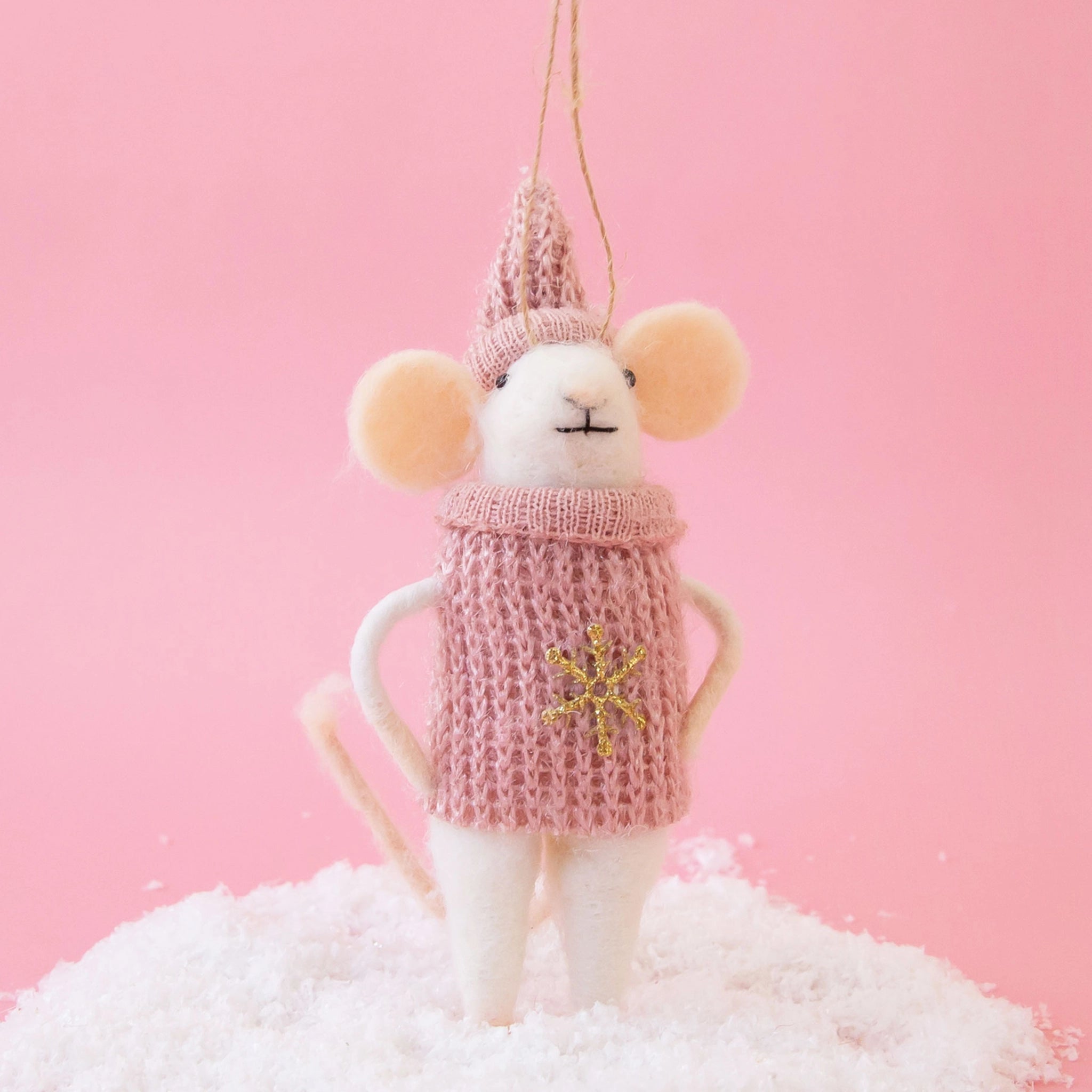 On a pink background is a white felt mouse ornament dressed in a pink knit sweater with a gold snowflake in the center and a pink matching hat. 