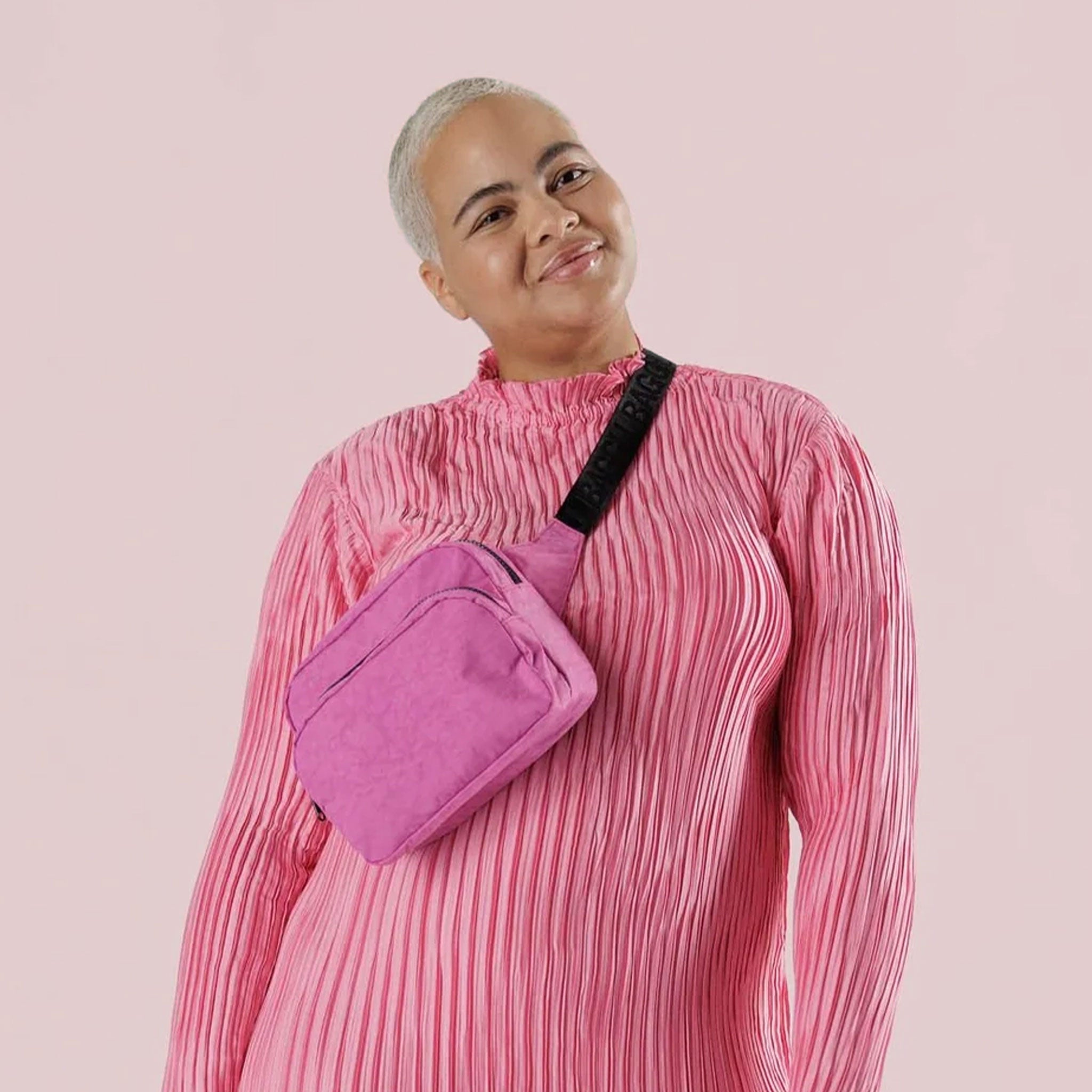 On a light pink background is a bright pink fanny pack with a black strap.
