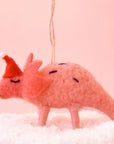 On a pink background is a coral colored felt dinosaur with a red santa hat on and standing on a pile of faux snow.