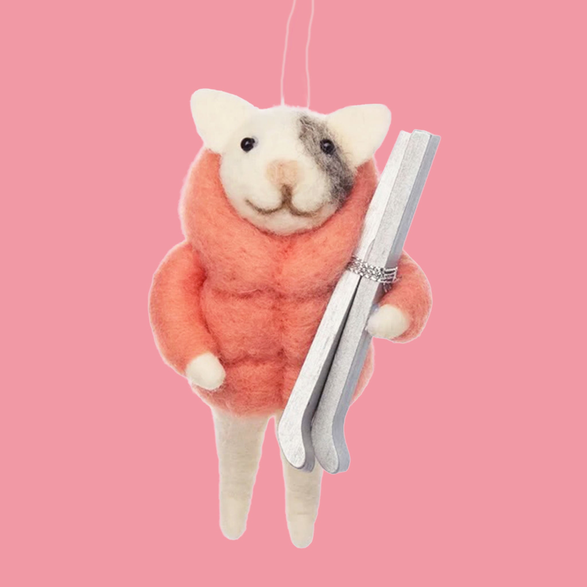On a pink background with a pink bristle brush tree is a white and grey felt cat ornament wearing a pink puffy jacket and holding a pair of silver skis. 