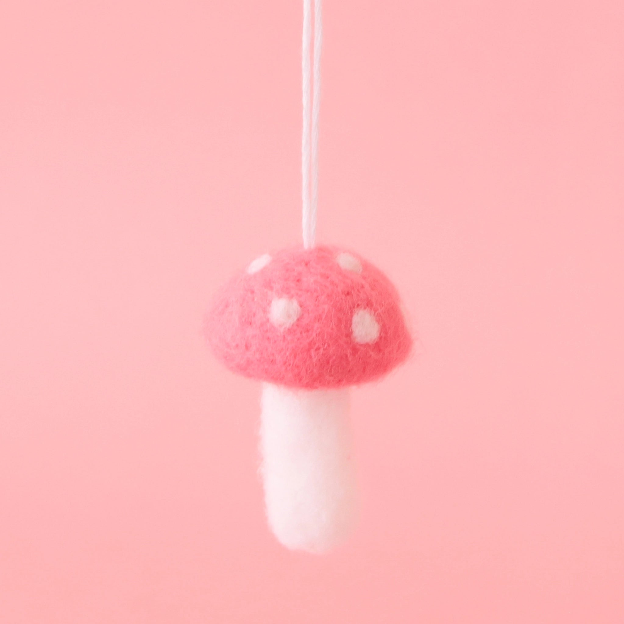 On a pink background is a dark pink felt ornament in the shape of a mushroom with white accents and dots. 
