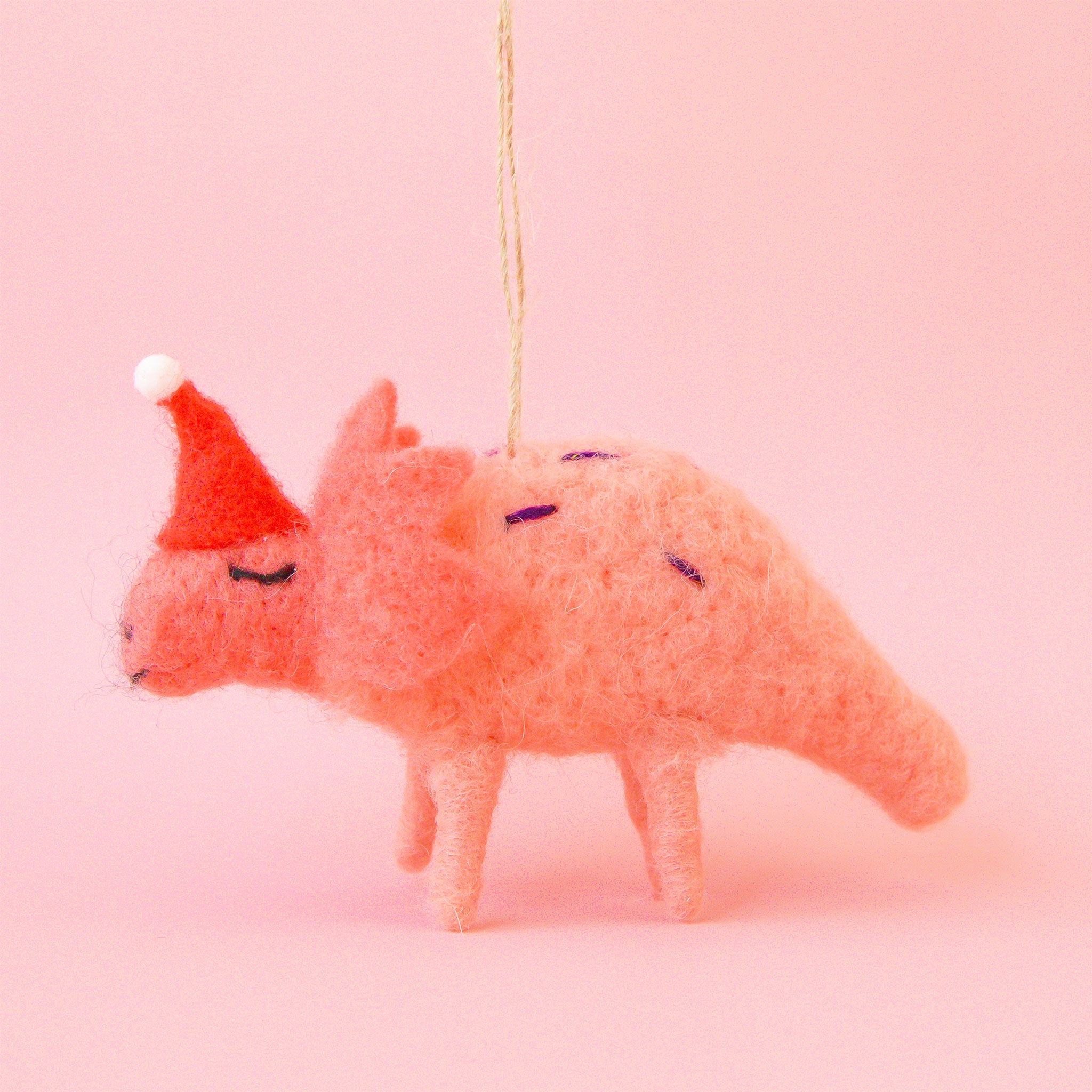 On a pink background is a coral colored felt dinosaur with a red santa hat.