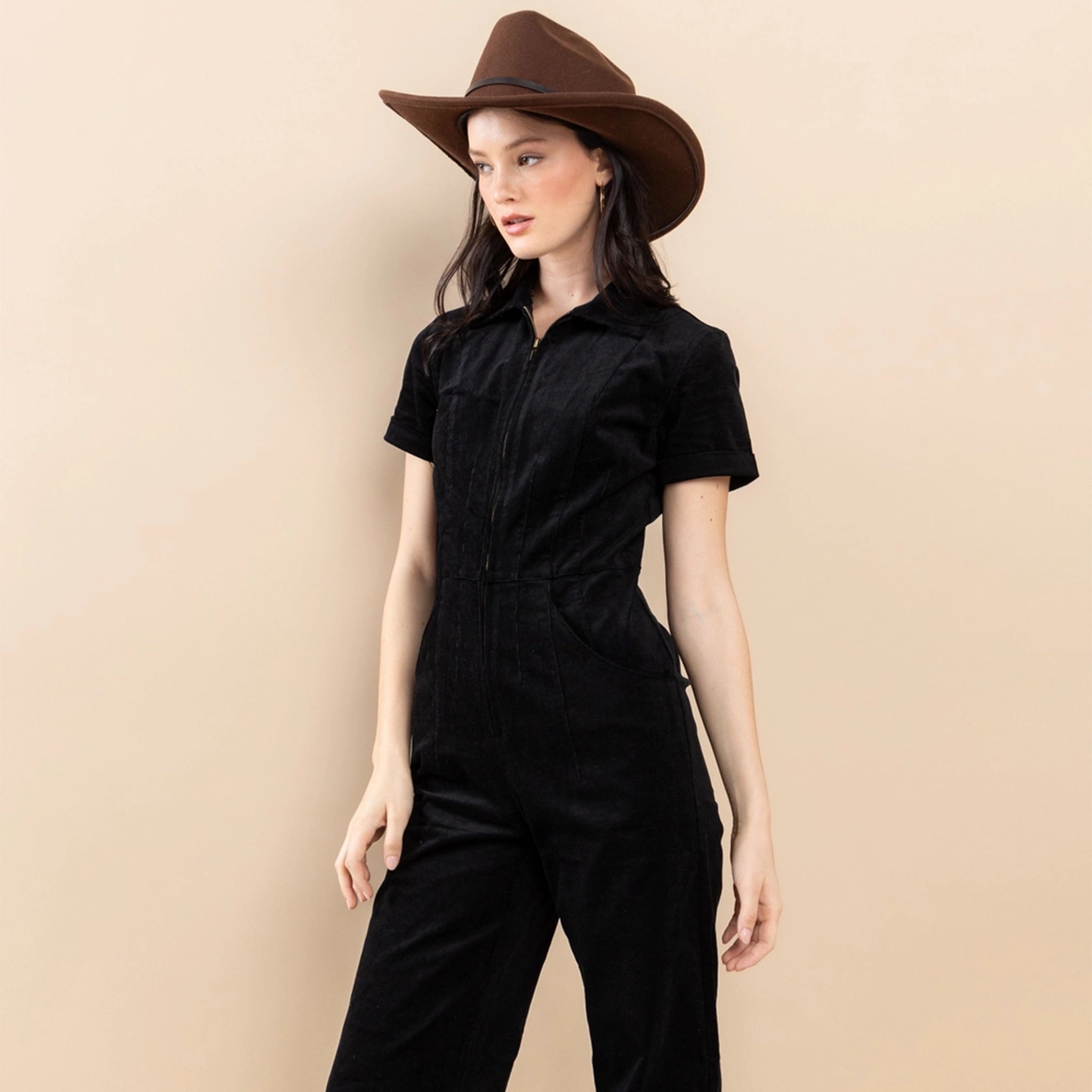 On a neutral background is a model wearing a black short sleeve jumpsuit with a front zipper. 