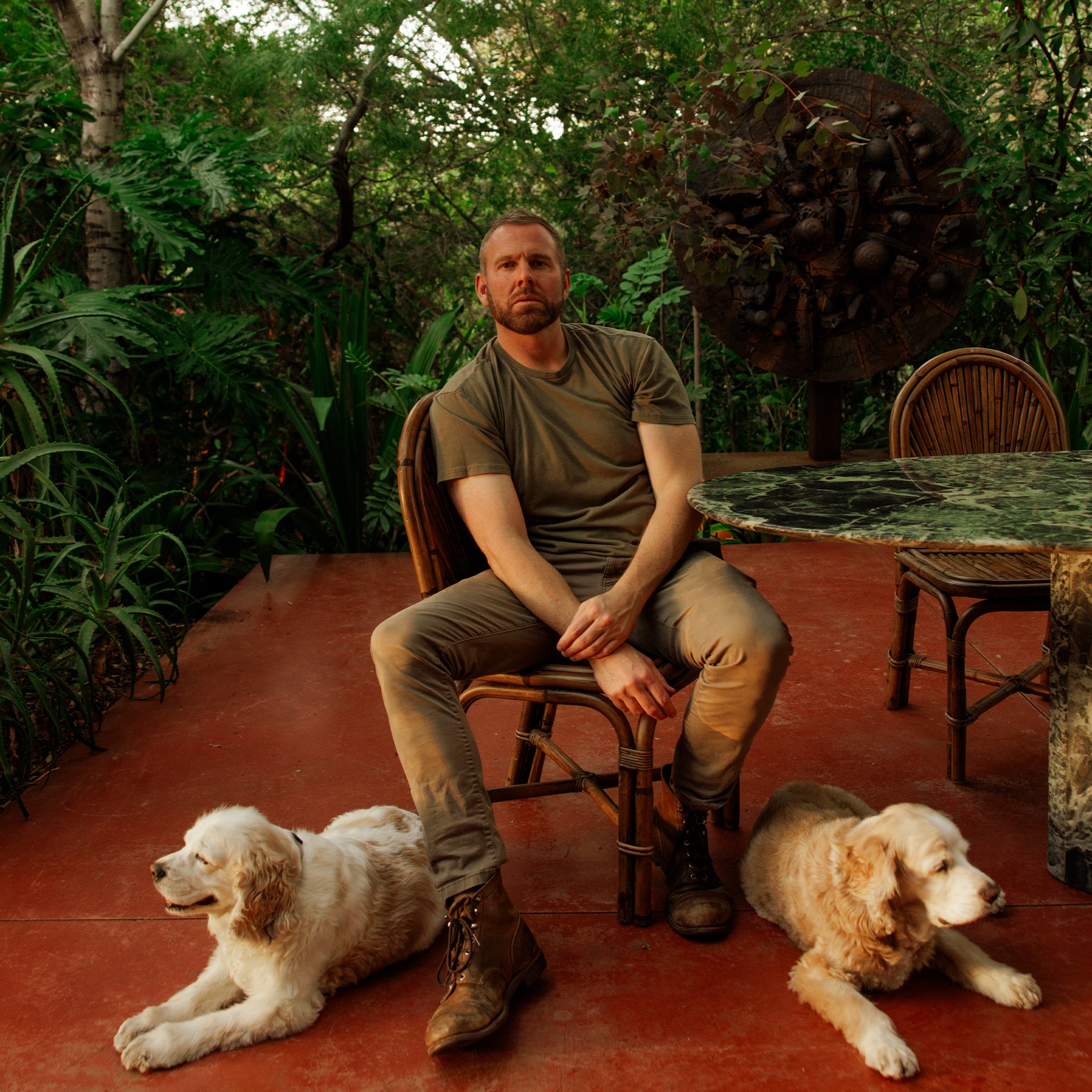 A man wearing an olive green t-shirt and brown pants sits on a wooden chair on a terracotta patio, flanked by two golden retriever dogs, with lush green foliage and a stone table in the background.