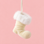A small Christmas boot shaped ornament in cream with a fluffy white faux-fur trim, hanging from a white ribbon against a solid pink background.