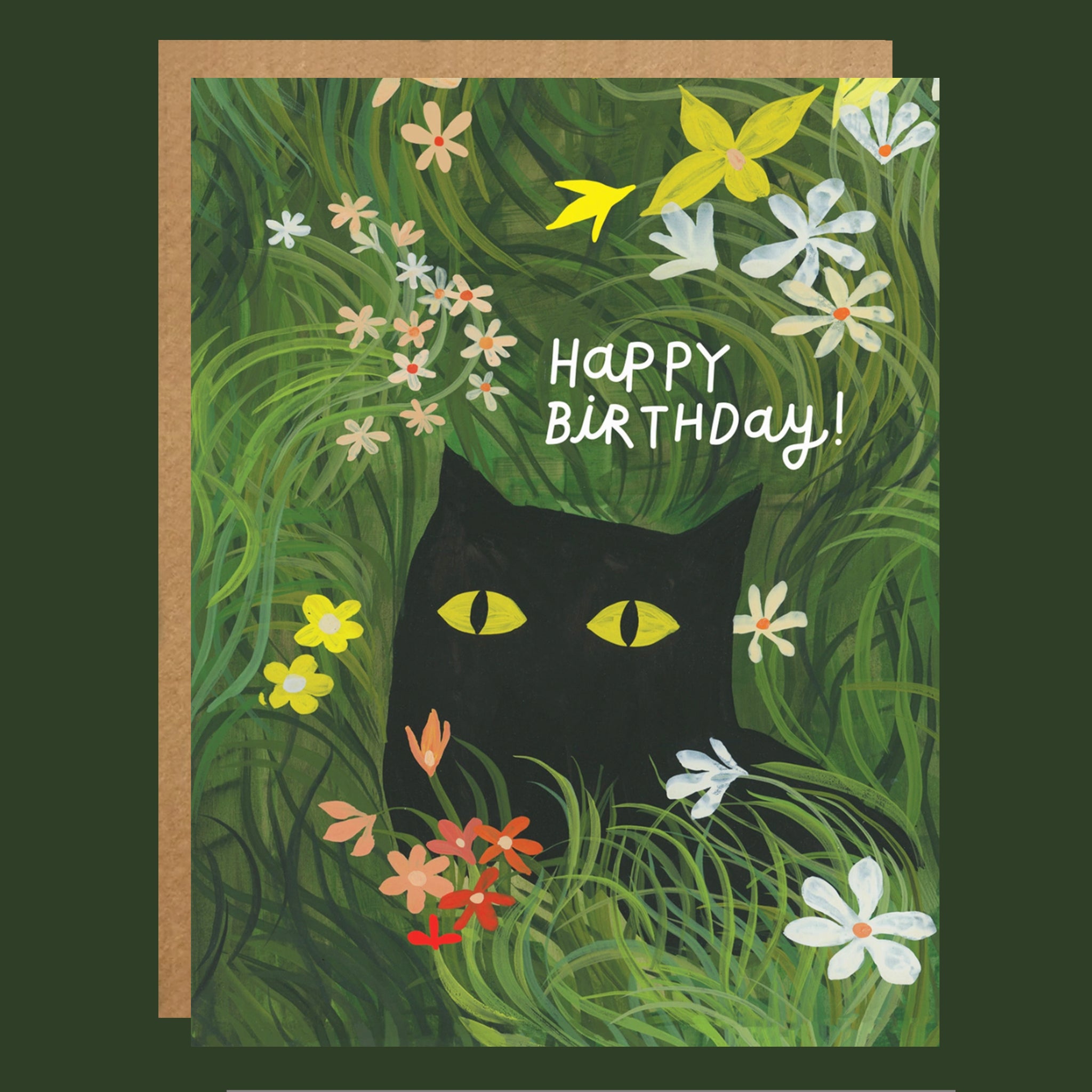 A square greeting card with a kraft envelope behind it, featuring a black cat with yellow eyes peeking through green grass among white, yellow, and pink flowers. The card reads 'Happy Birthday!' in white lettering.