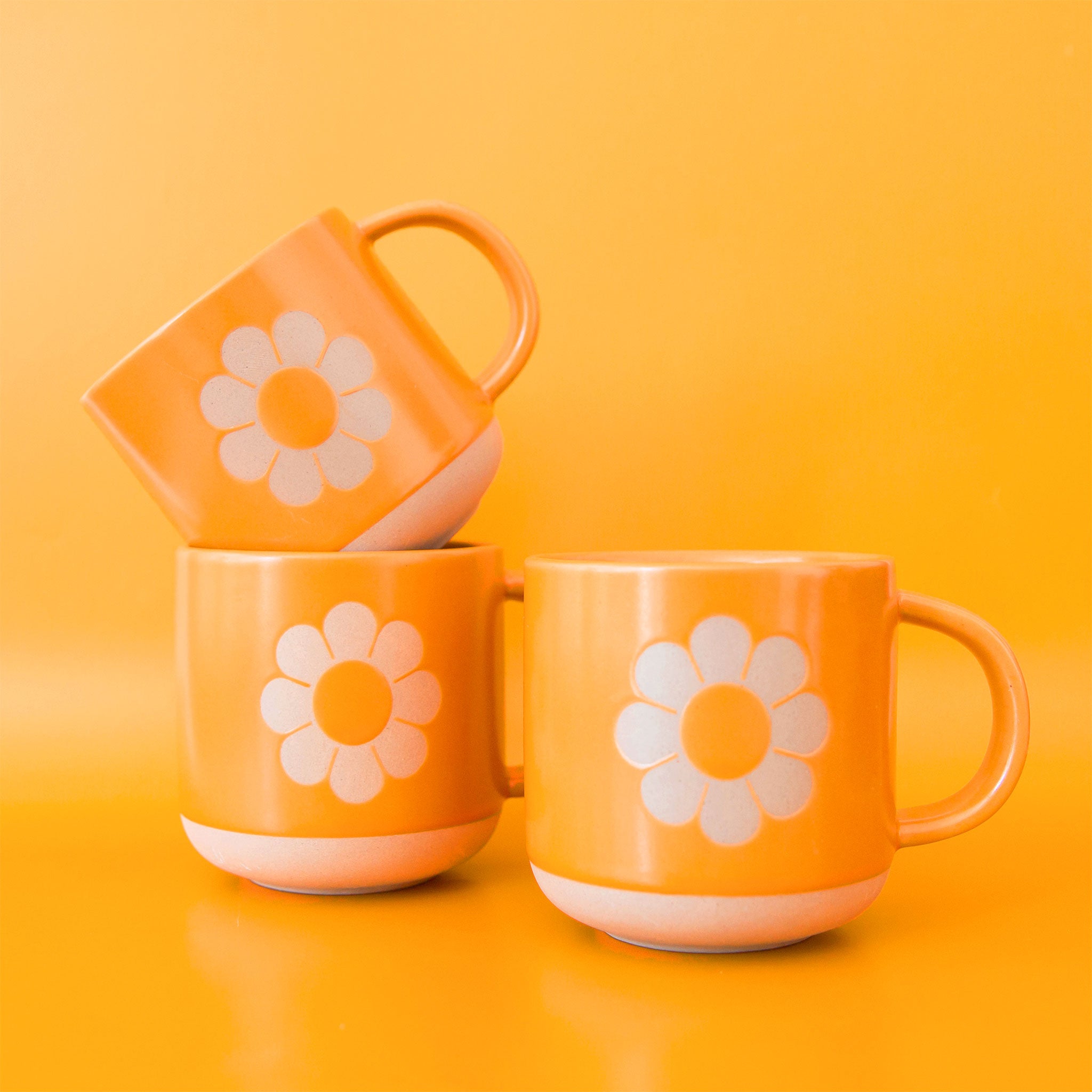 On a bright yellow background is three ceramic mugs with a daisy design on the front. 