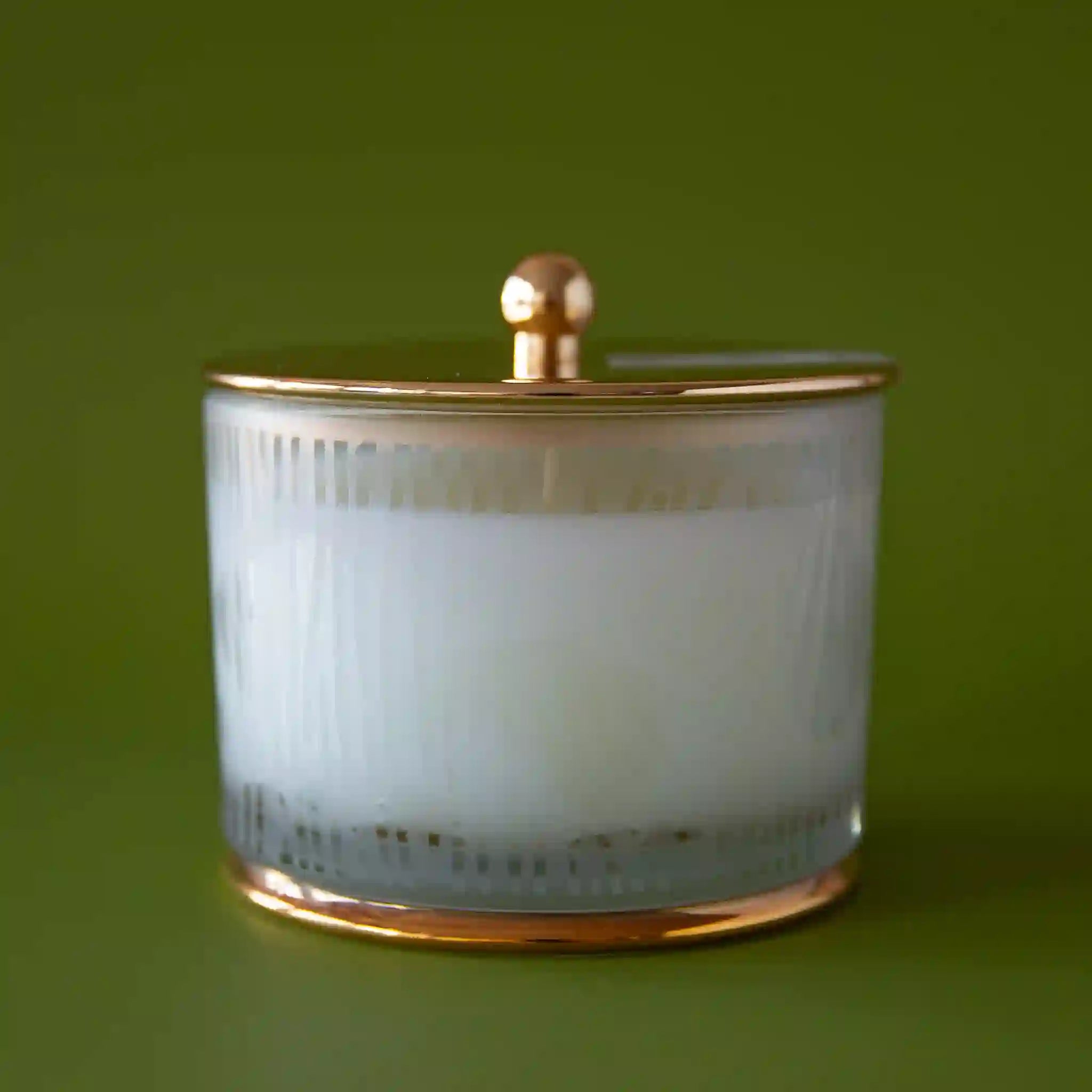 On a dark green background is a frosted glass jar candle with a gold lid.
