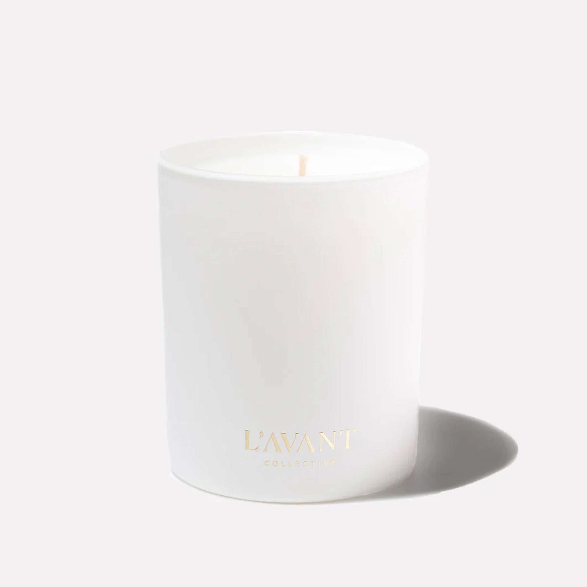A white candle with gold writing that reads, "L'avant Collective" on a white background.