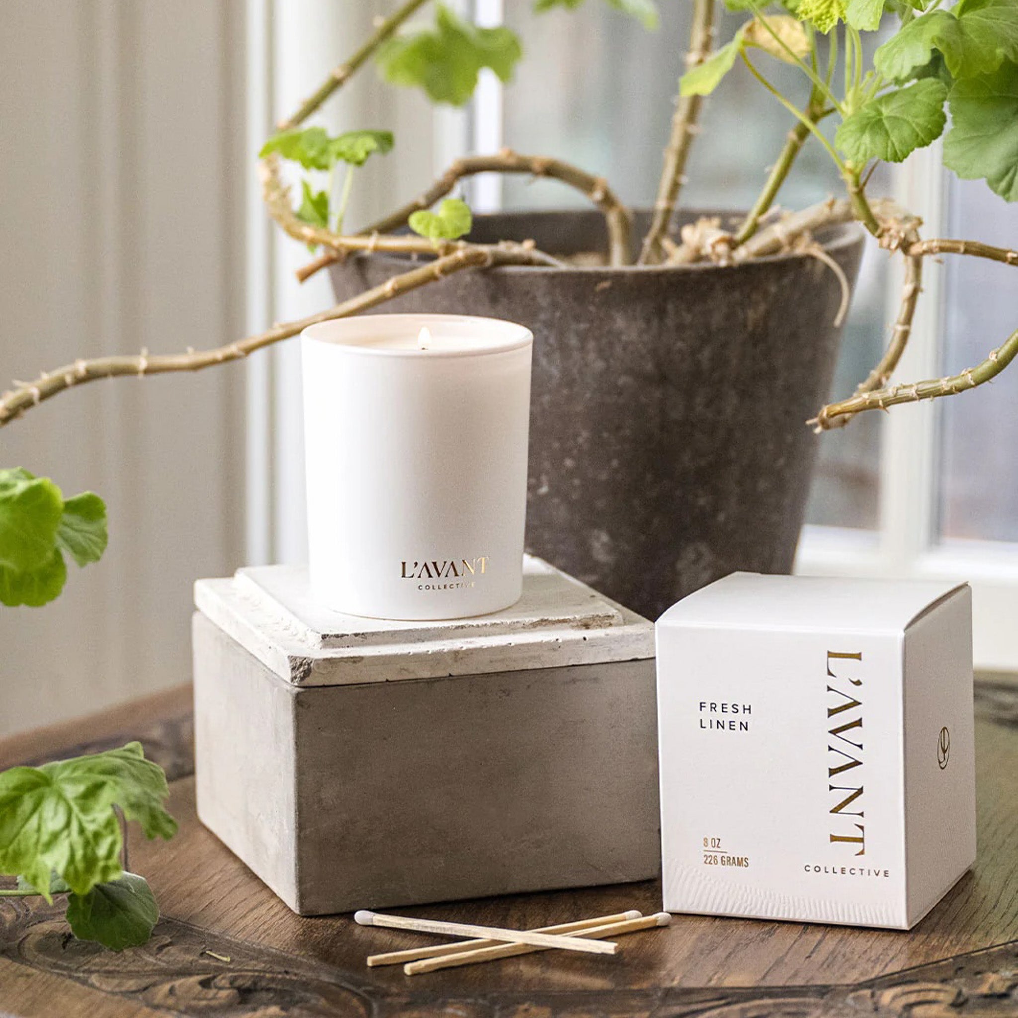 L’AVANT Collective Fresh Linen scented candle on a marble block beside its box and matches, styled near a potted plant.